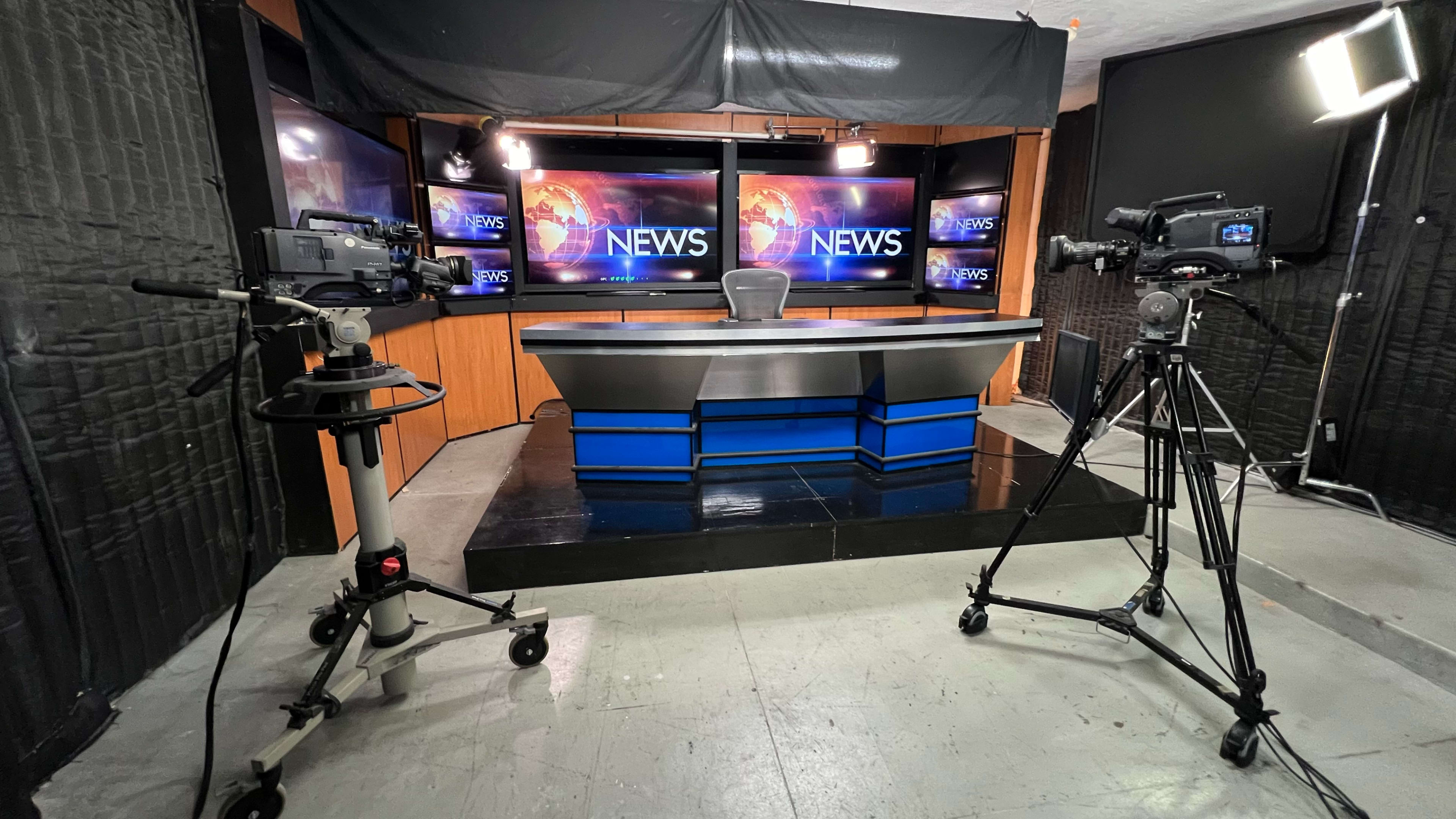 The image shows a news studio setup with a central news desk, multiple cameras, and large screens displaying the word "NEWS."
