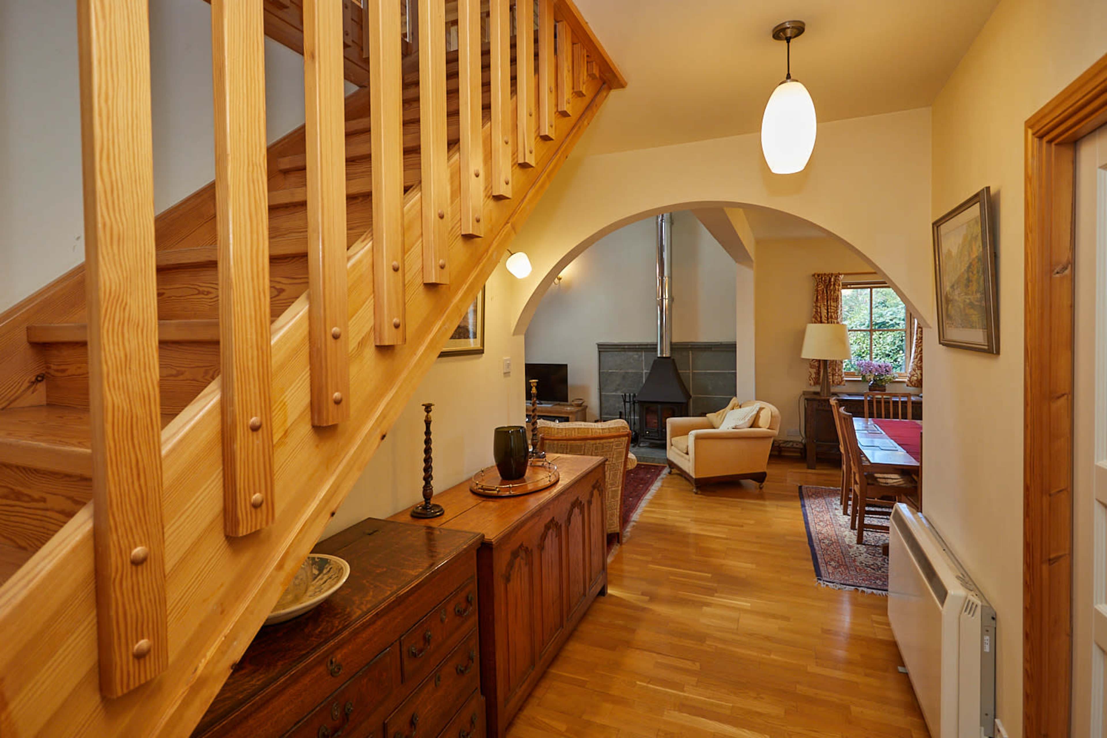 A wooden staircase leads from a hallway into a living area with a fireplace, flanked by a dining table and antique furniture.