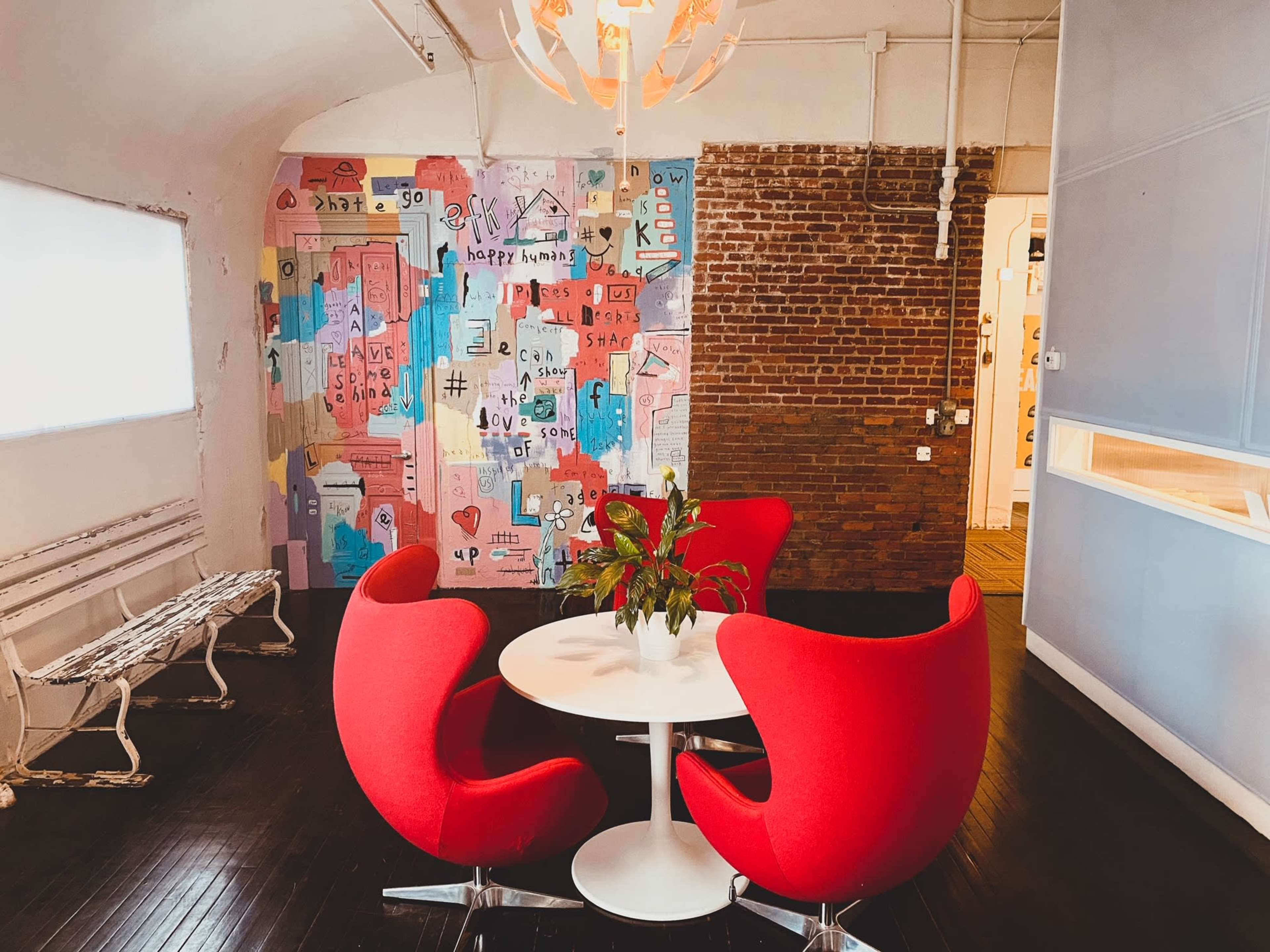 A small dining area features a round white table surrounded by three red chairs, with a colorful mural on the wall and a plant at the center of the table.