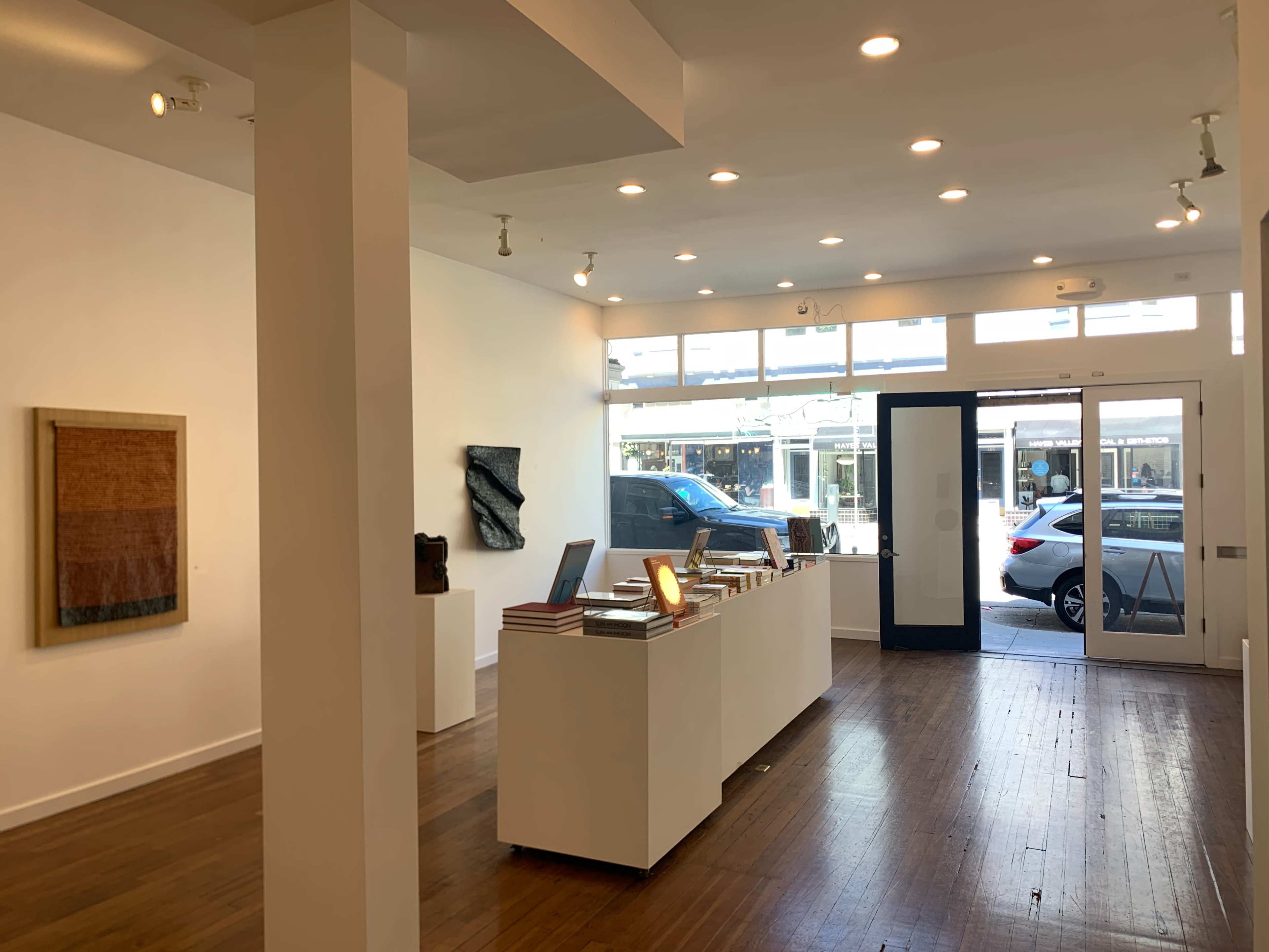 The image shows a bright, minimalist art gallery with wooden flooring and large front windows displaying artworks and a view of parked cars outside.