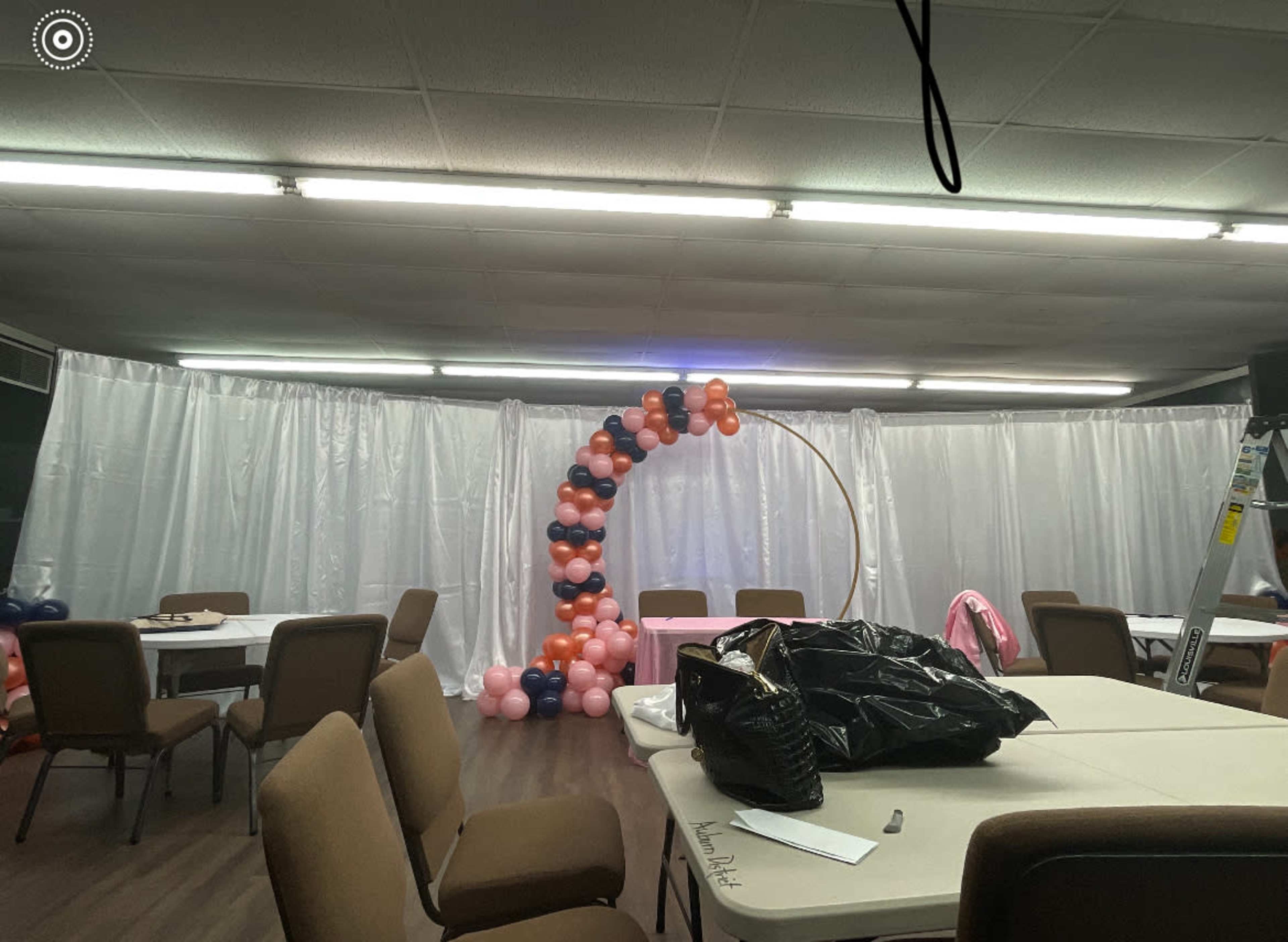 A partially set up event space featuring a circular balloon arch and several tables surrounded by chairs.