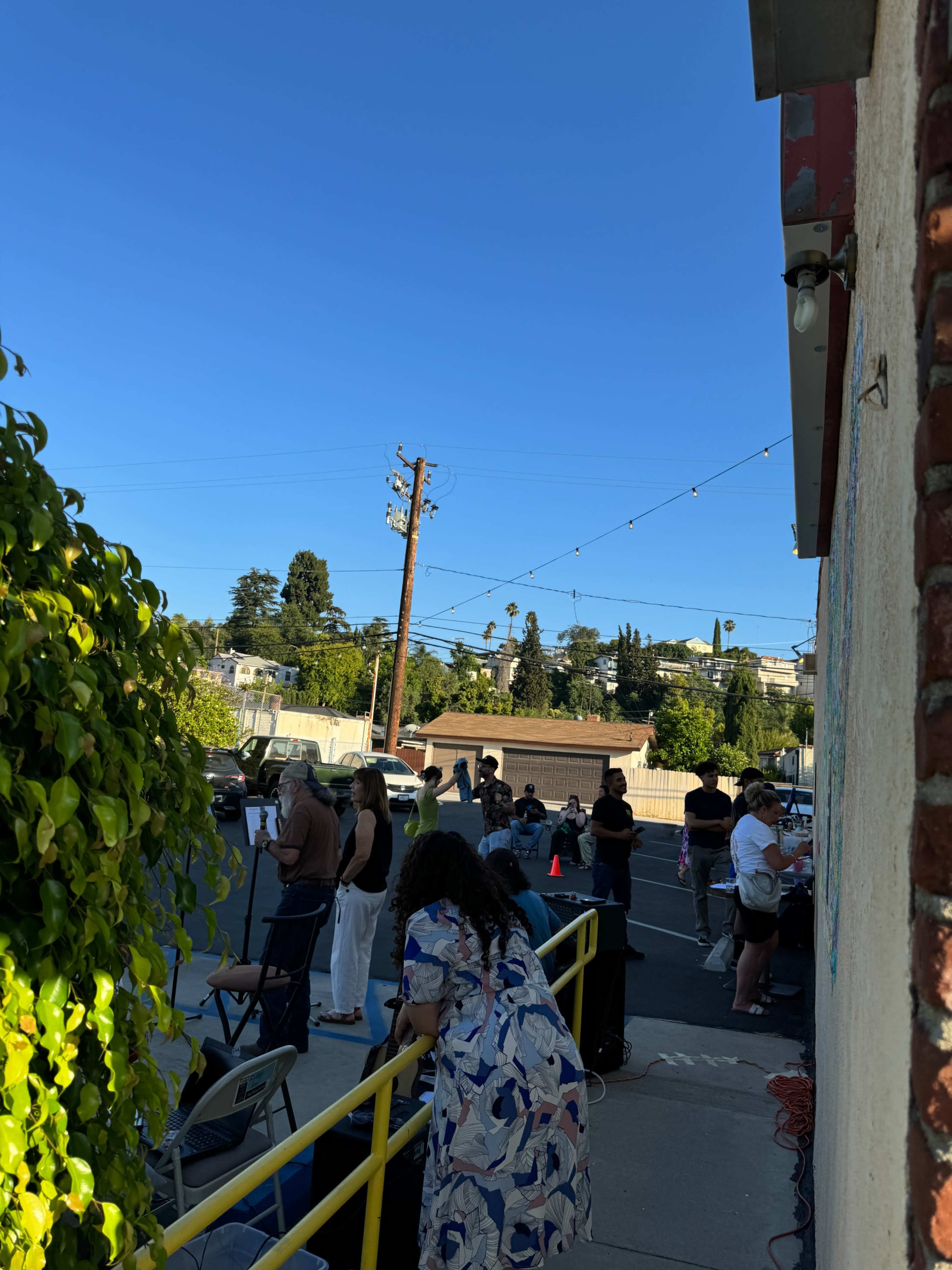 People gather outside a building on a sunny day, surrounded by parked cars and greenery.
