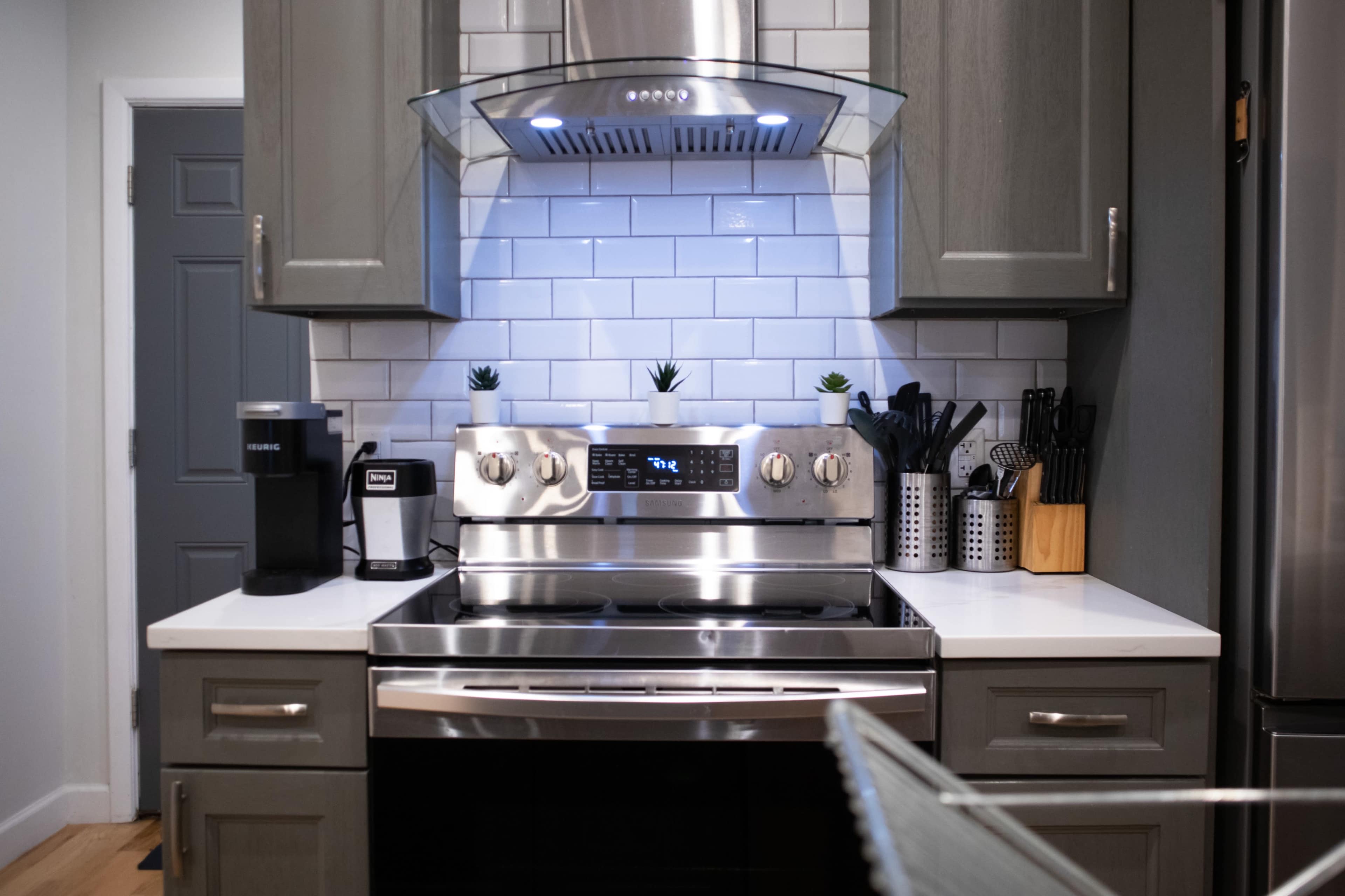 The image shows a modern kitchen with a stainless steel stove, hood, and various kitchen utensils organized on the countertop.