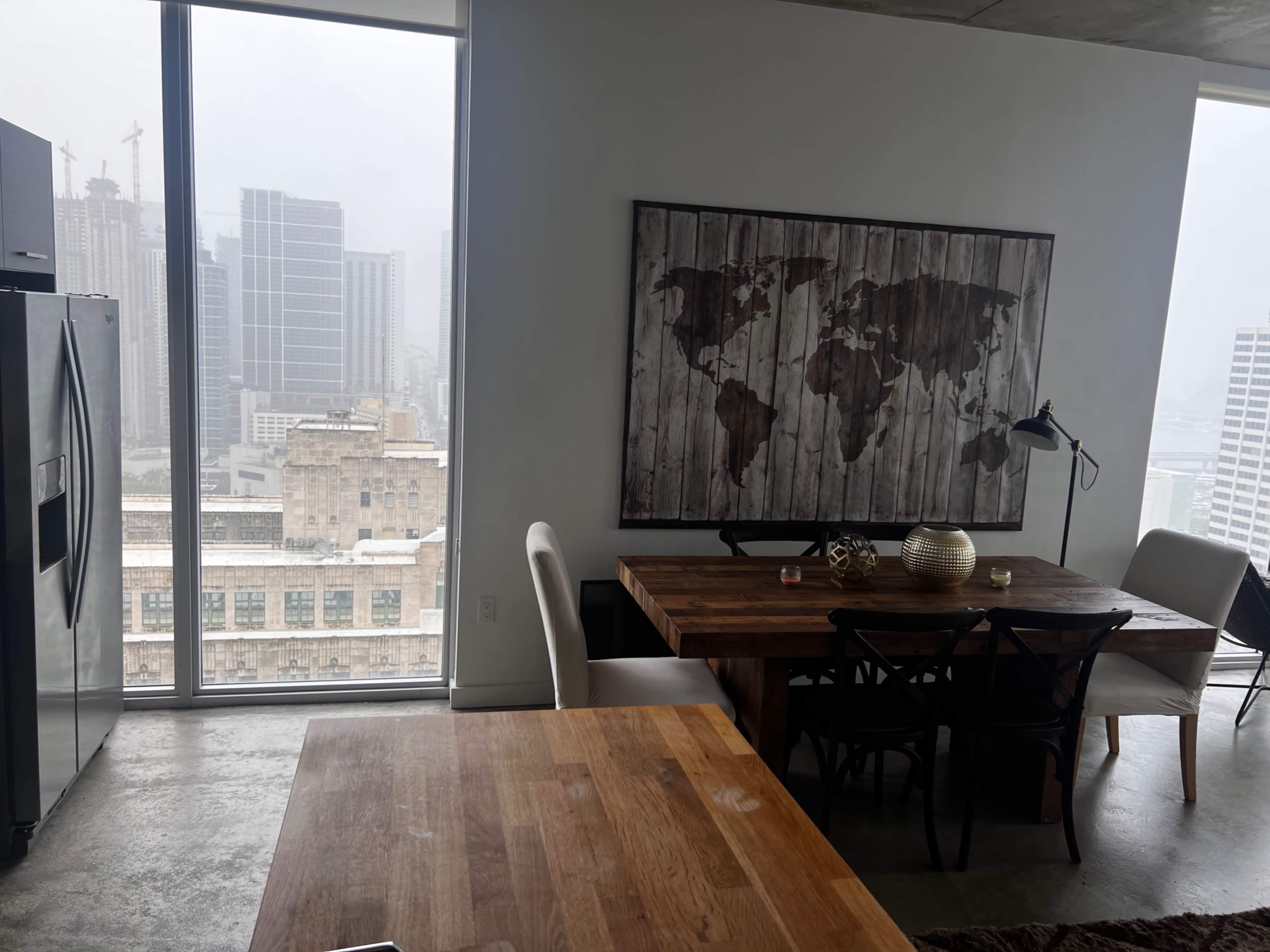 The image shows a modern kitchen and dining area with a wooden table and a large world map artwork on the wall, overlooking a city skyline through tall windows.