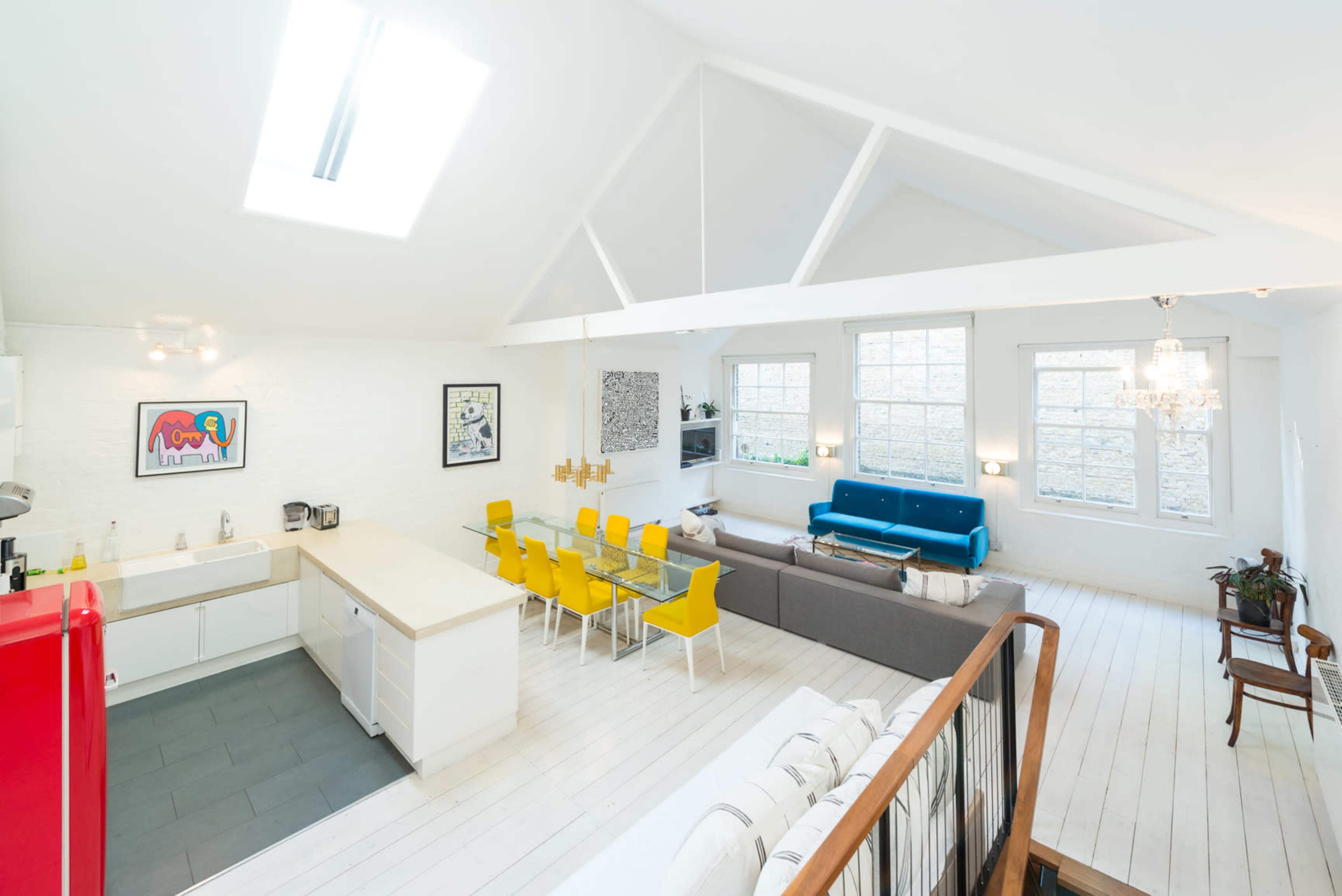 The image shows a bright, open-concept living space featuring a kitchen, dining area with a yellow table and chairs, and a lounge area with blue and gray sofas.