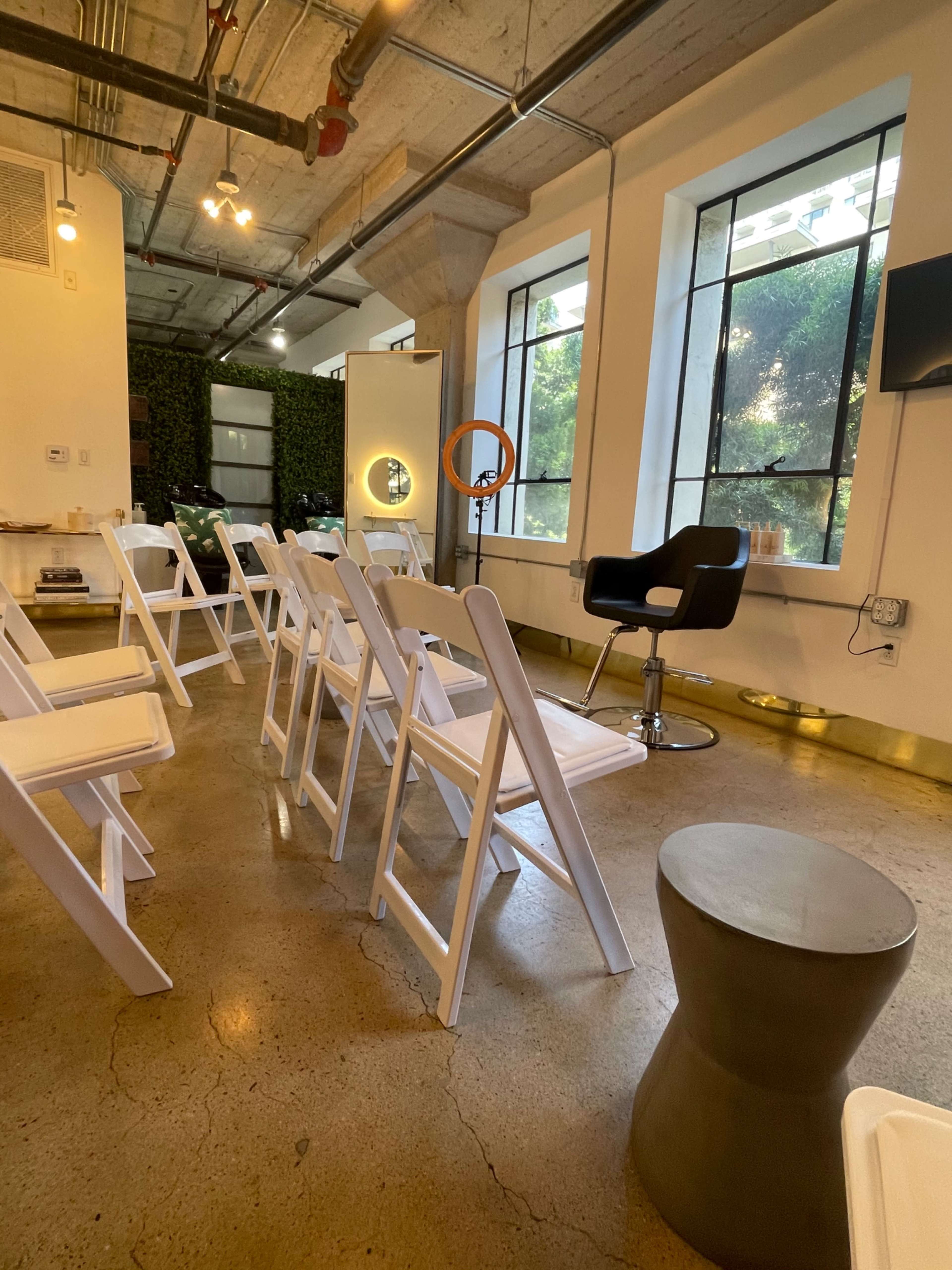 The image shows a spacious room set up with white folding chairs facing a styling chair and a ring light near large windows.