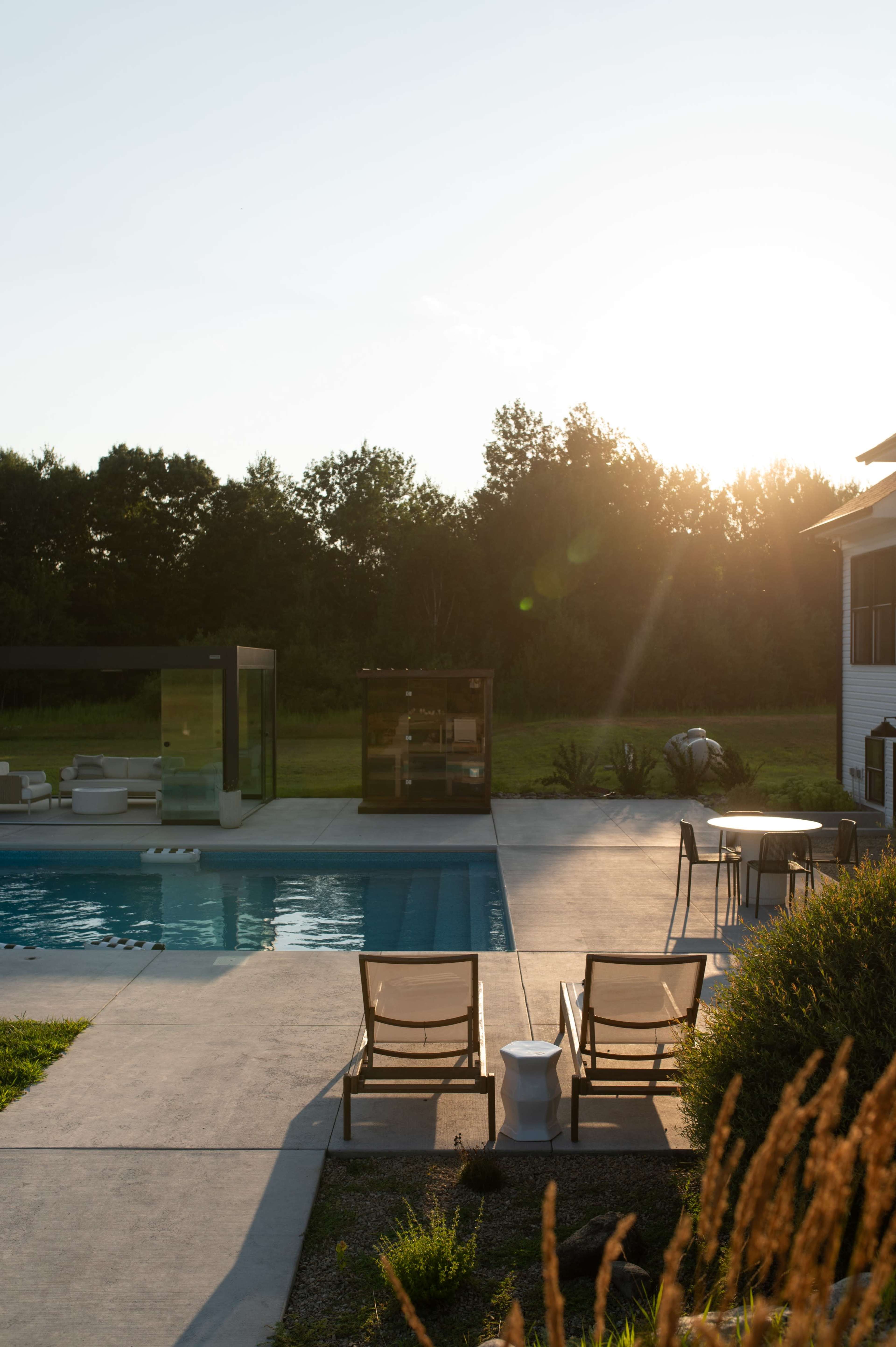 The image shows a modern backyard with a swimming pool, lounge chairs, and a small structure surrounded by green foliage under a sunset sky.