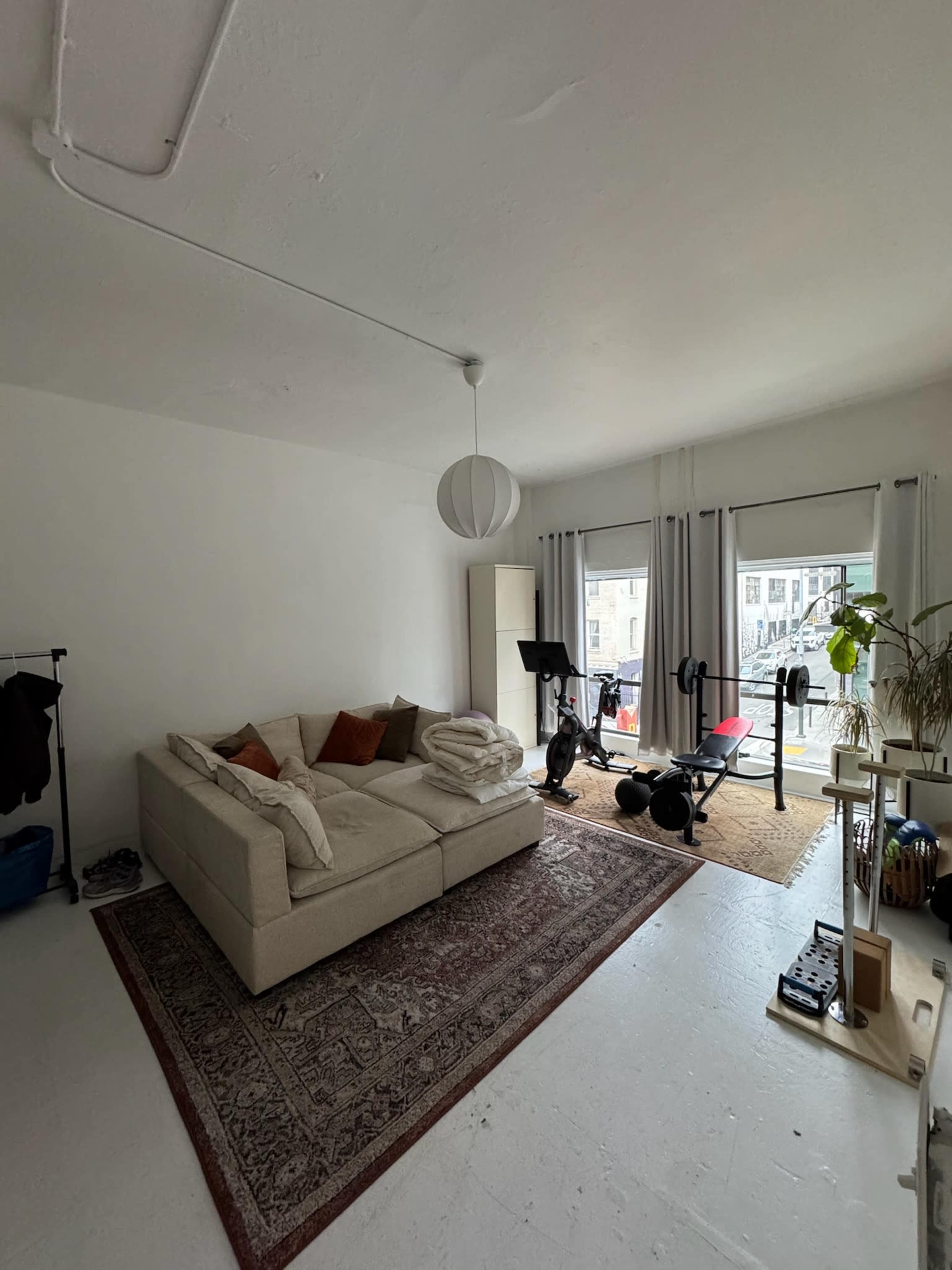 The image shows a sparsely furnished living room featuring a sofa, a rug, and a workout area with gym equipment near a window.