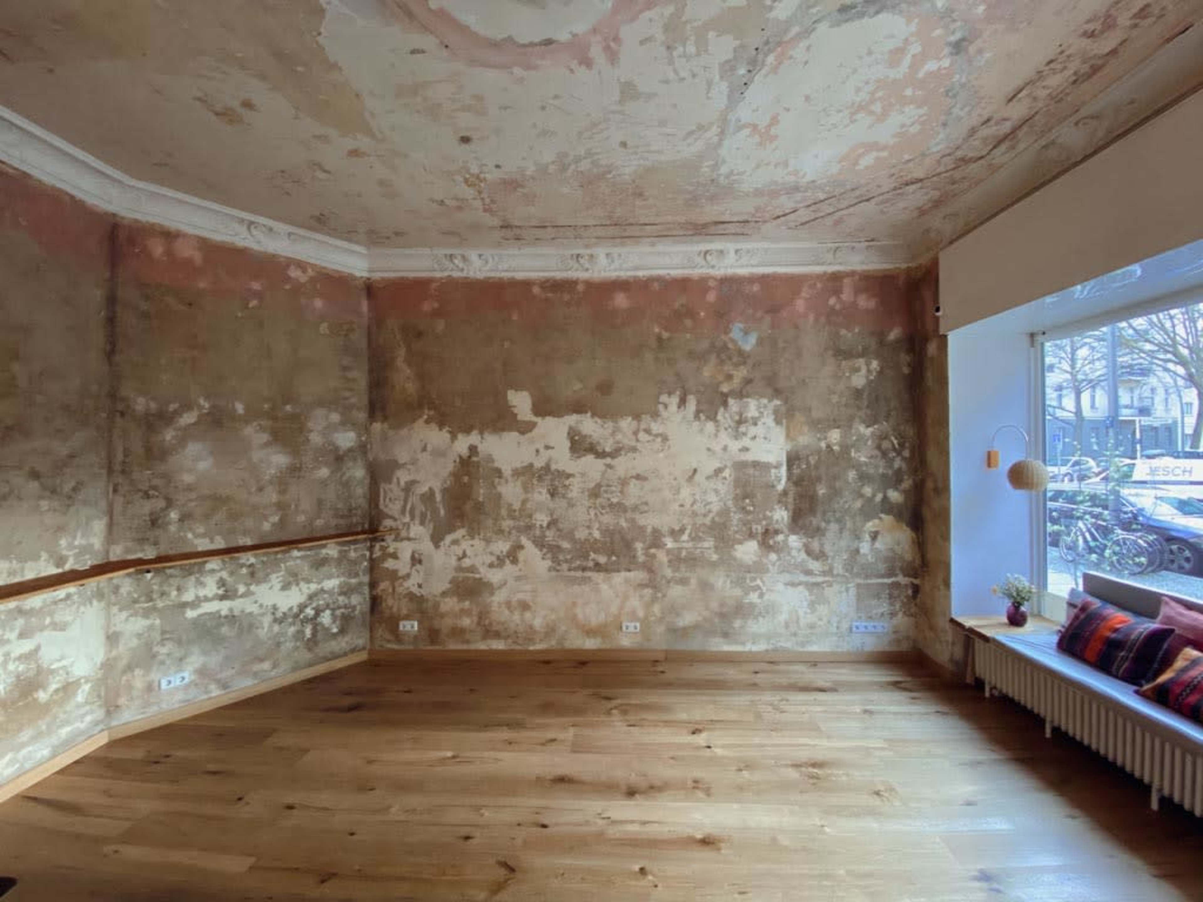 The image shows an empty room with peeling walls, wooden flooring, and a large window revealing a view of bicycles outside.