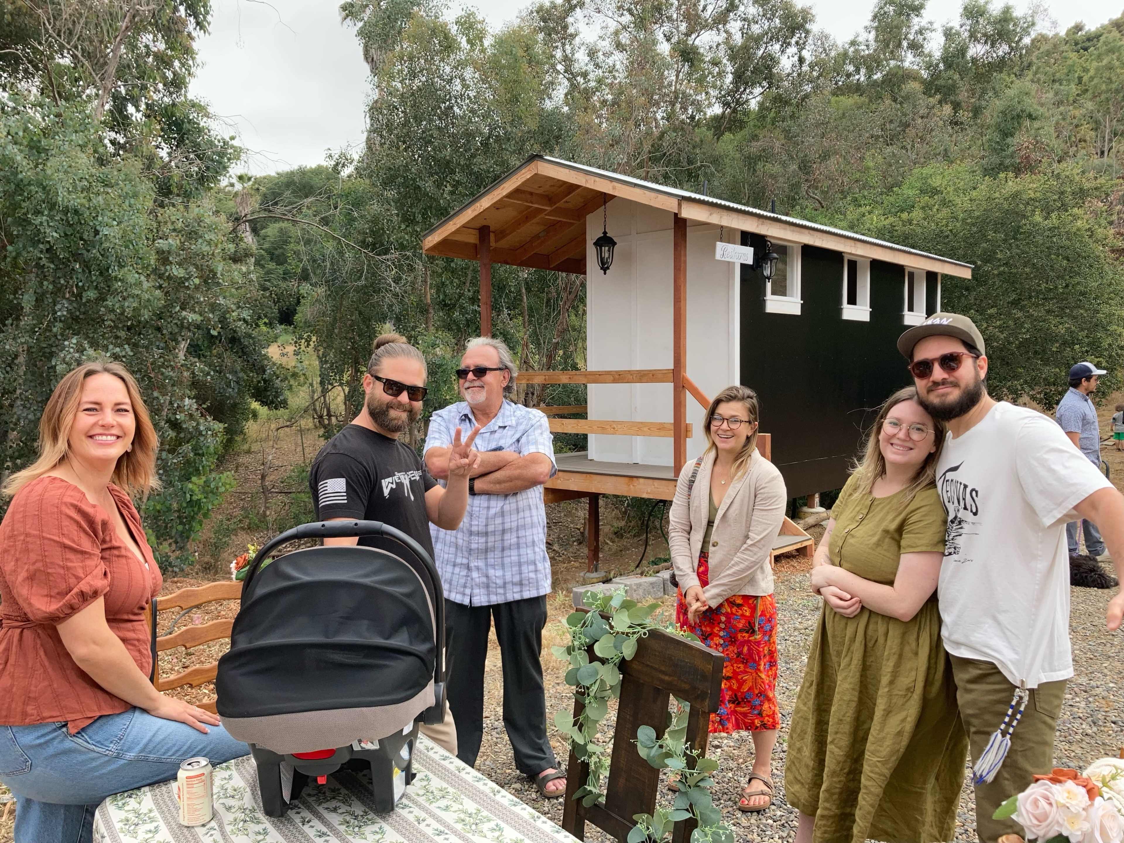 A group of six people stands in front of a small black and white cabin, surrounded by trees and outdoor furniture.