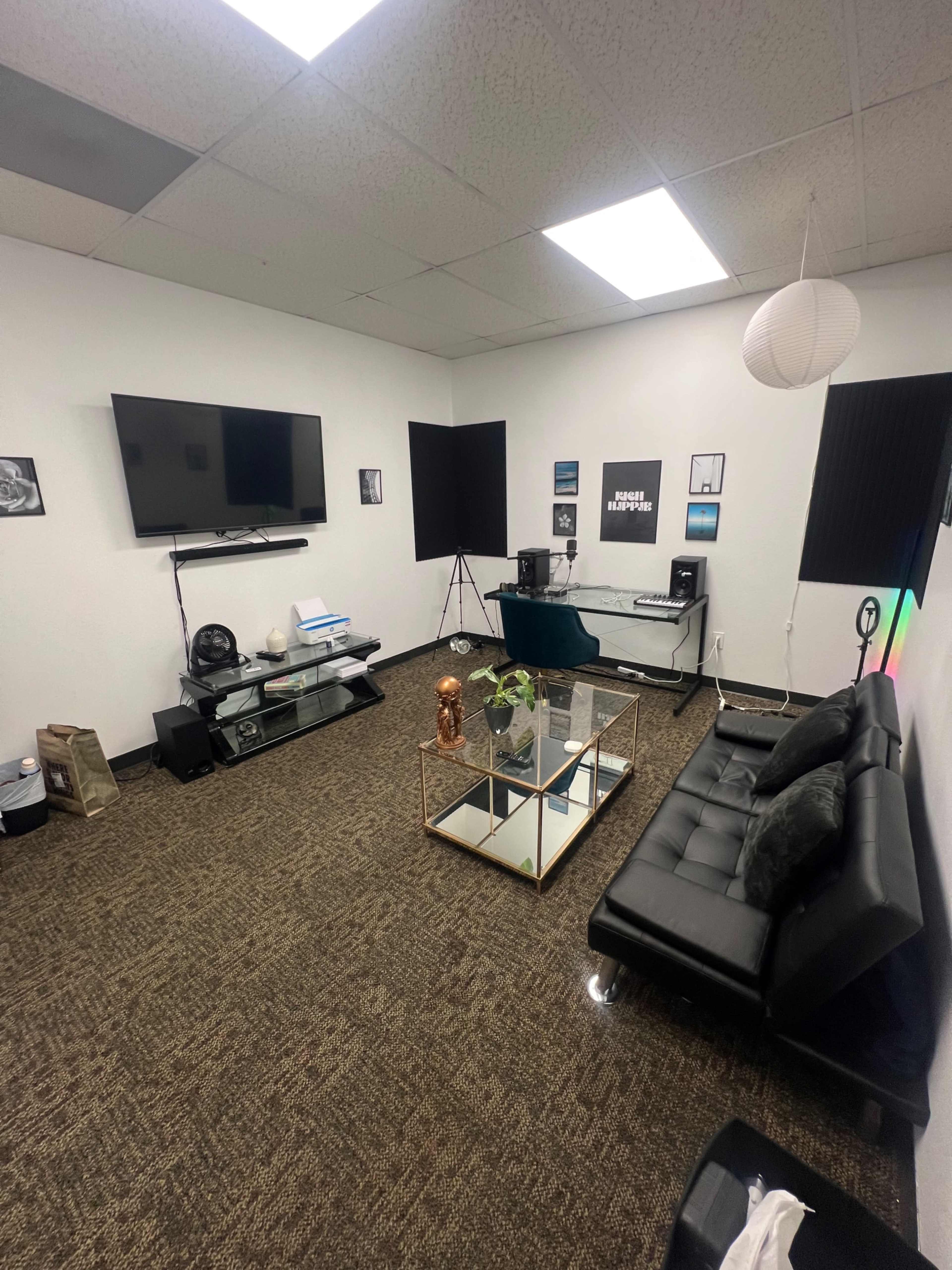 The image shows a neatly arranged room featuring a black sofa, a glass coffee table, a TV mounted on the wall, and a music setup in one corner.