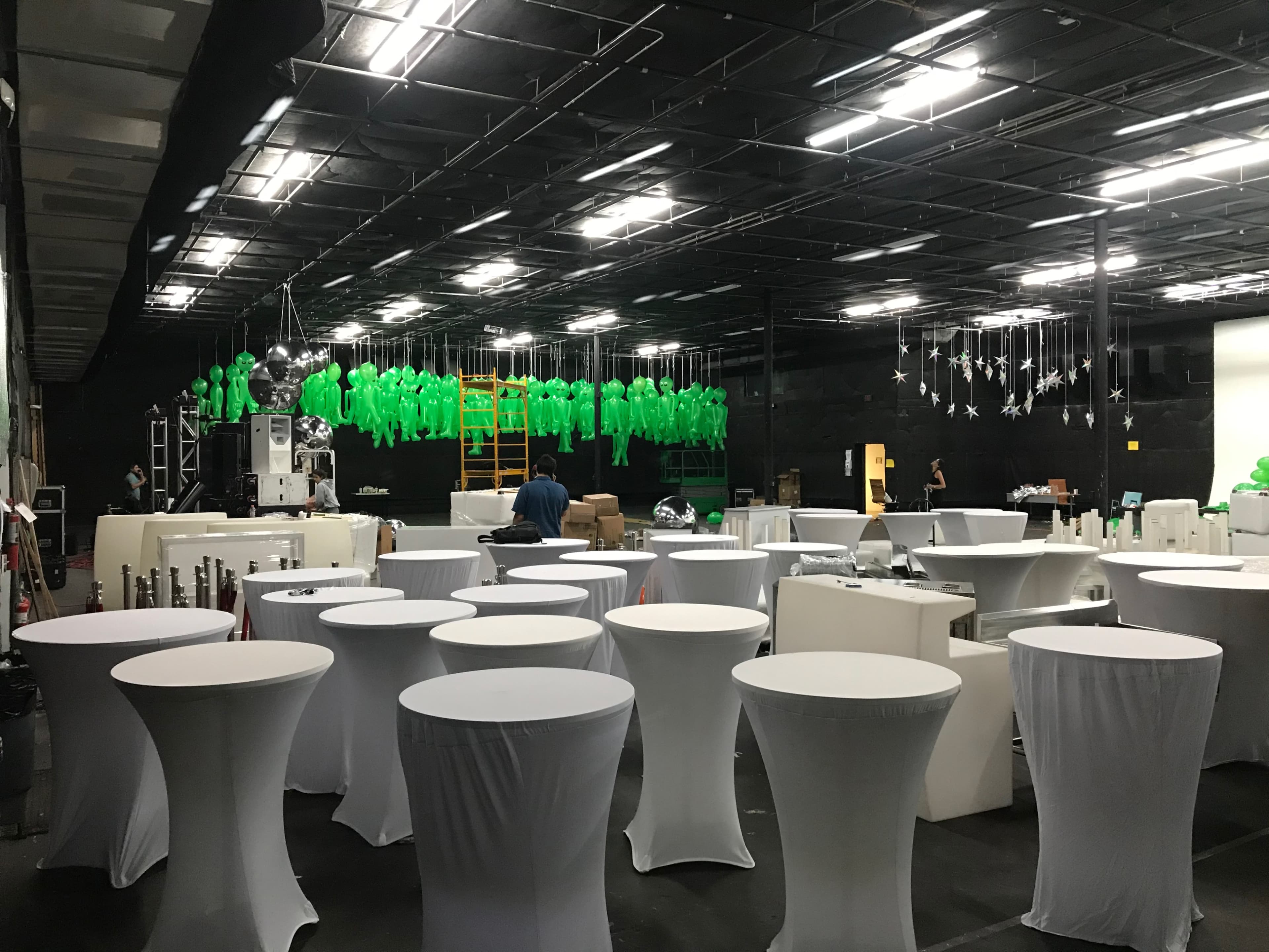 The image shows a large event space with white tables surrounded by various decorations, including green figures hanging from the ceiling.