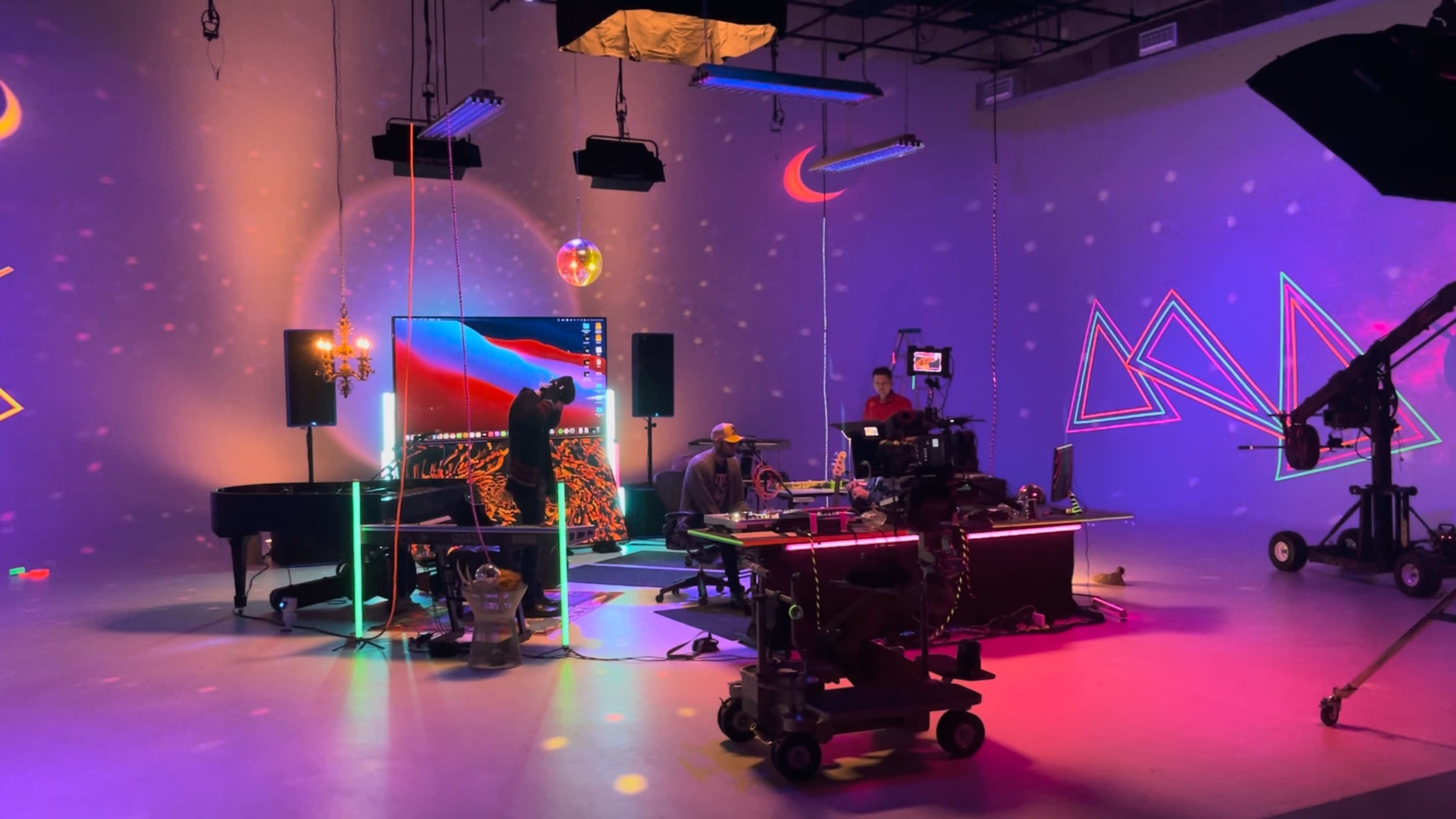 The image shows a vibrant studio setup with colorful lighting, a large screen displaying graphics, and several people working around a central table with film equipment.