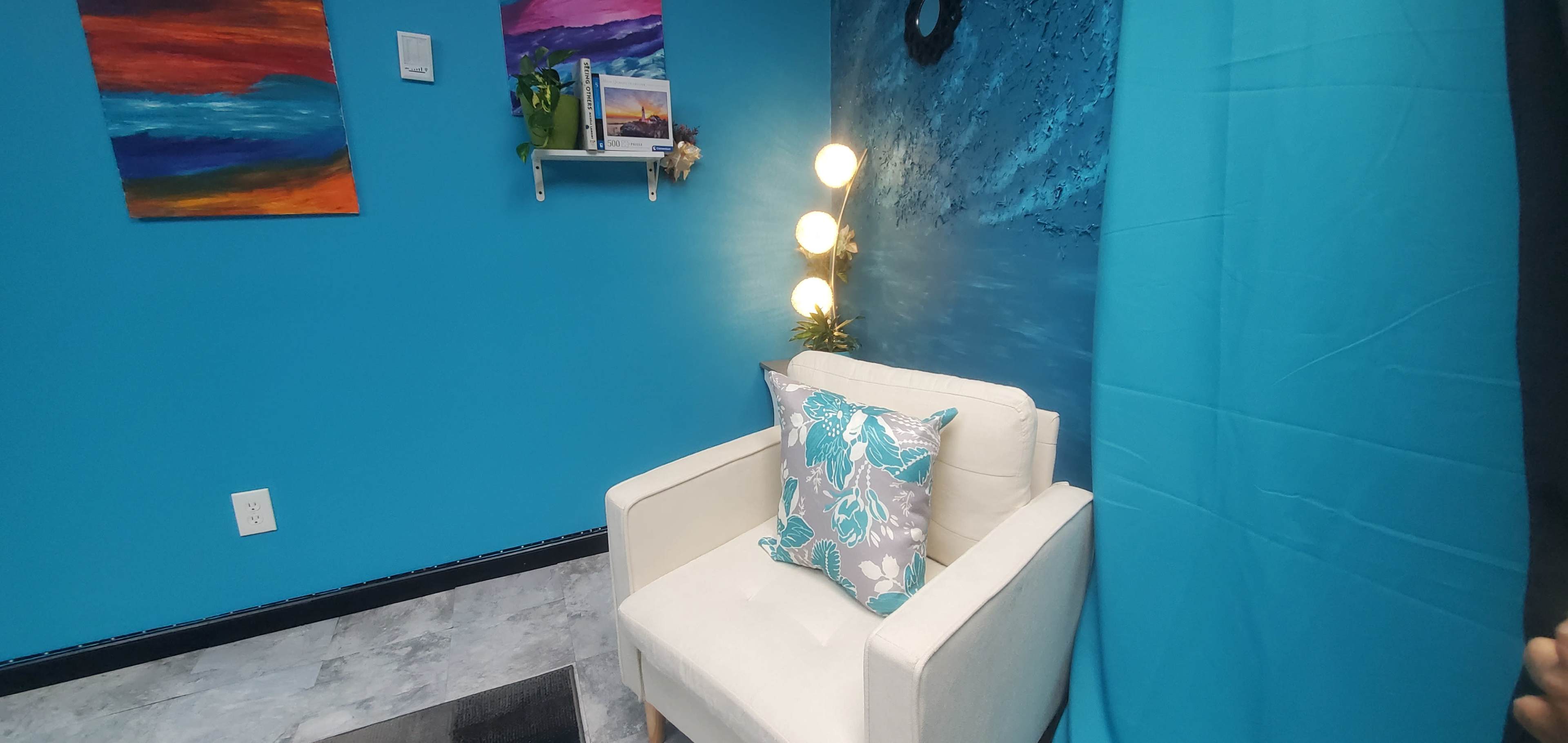 A brightly colored room with turquoise walls and a white chair adorned with a floral cushion, next to a small shelf holding decorative plants and artwork.