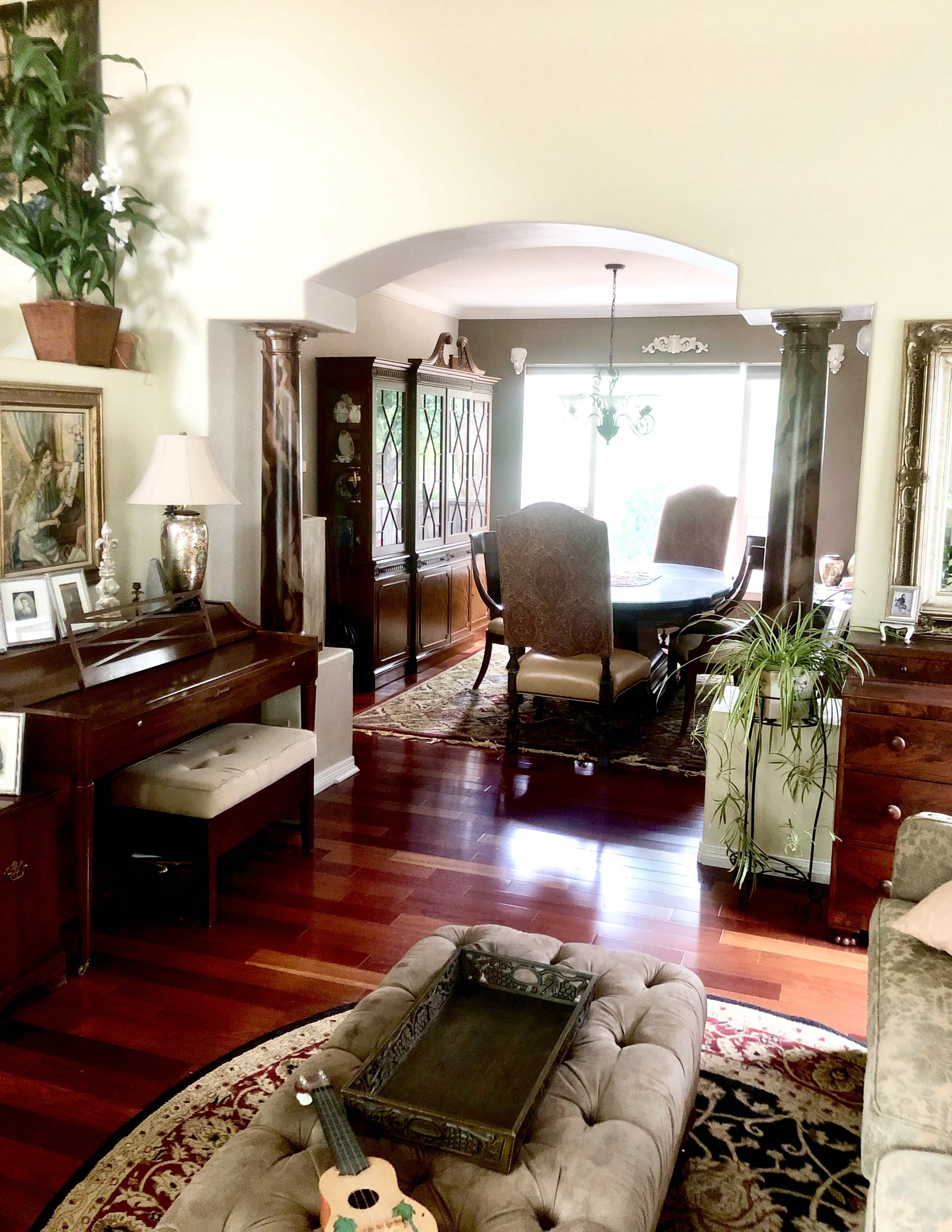 A living room featuring wooden furniture, a piano, and a view into a dining area with a large table and ornate decor.