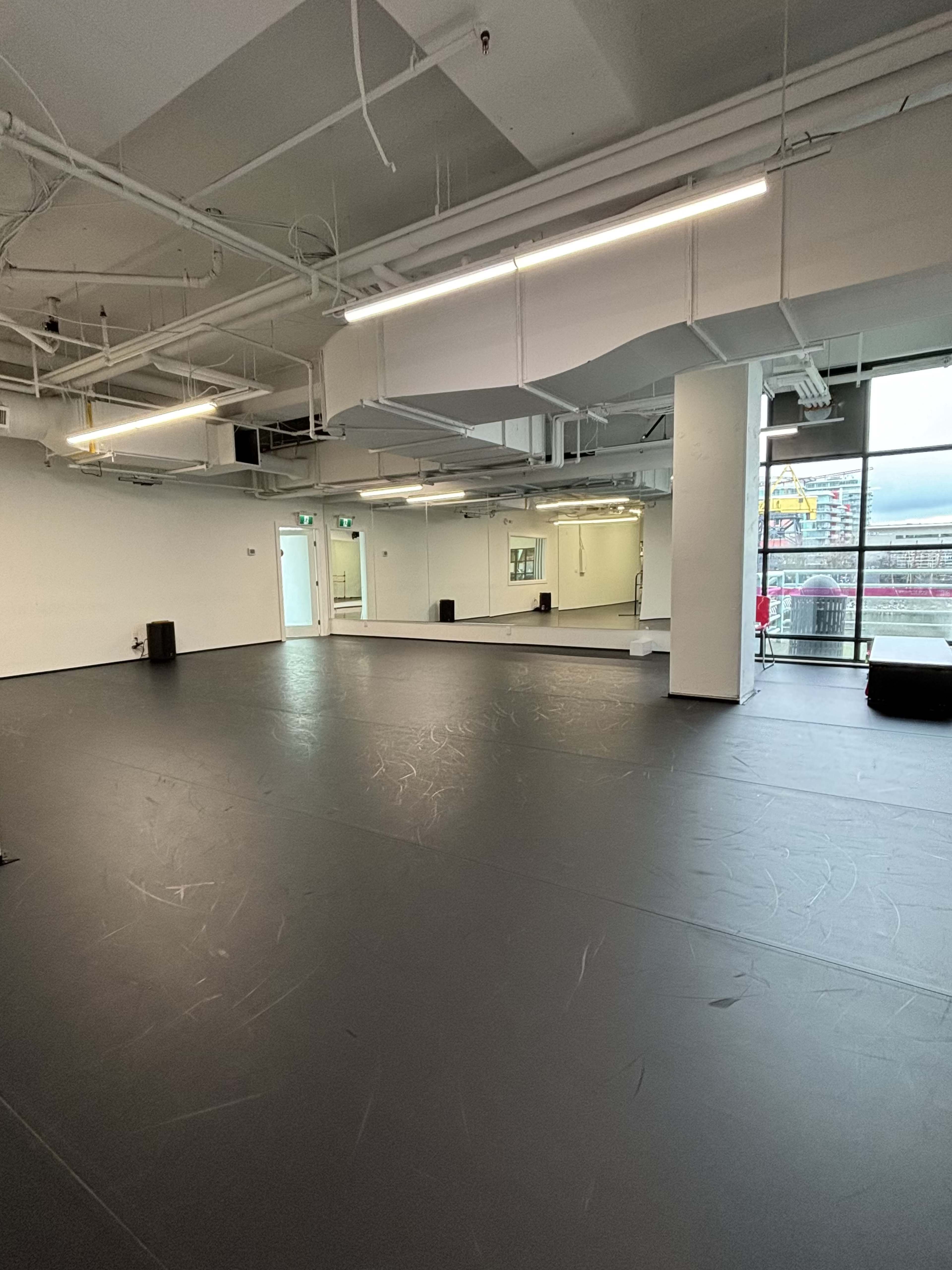 The image shows a large, empty studio space with black flooring, bright overhead lights, and a wall of mirrors.