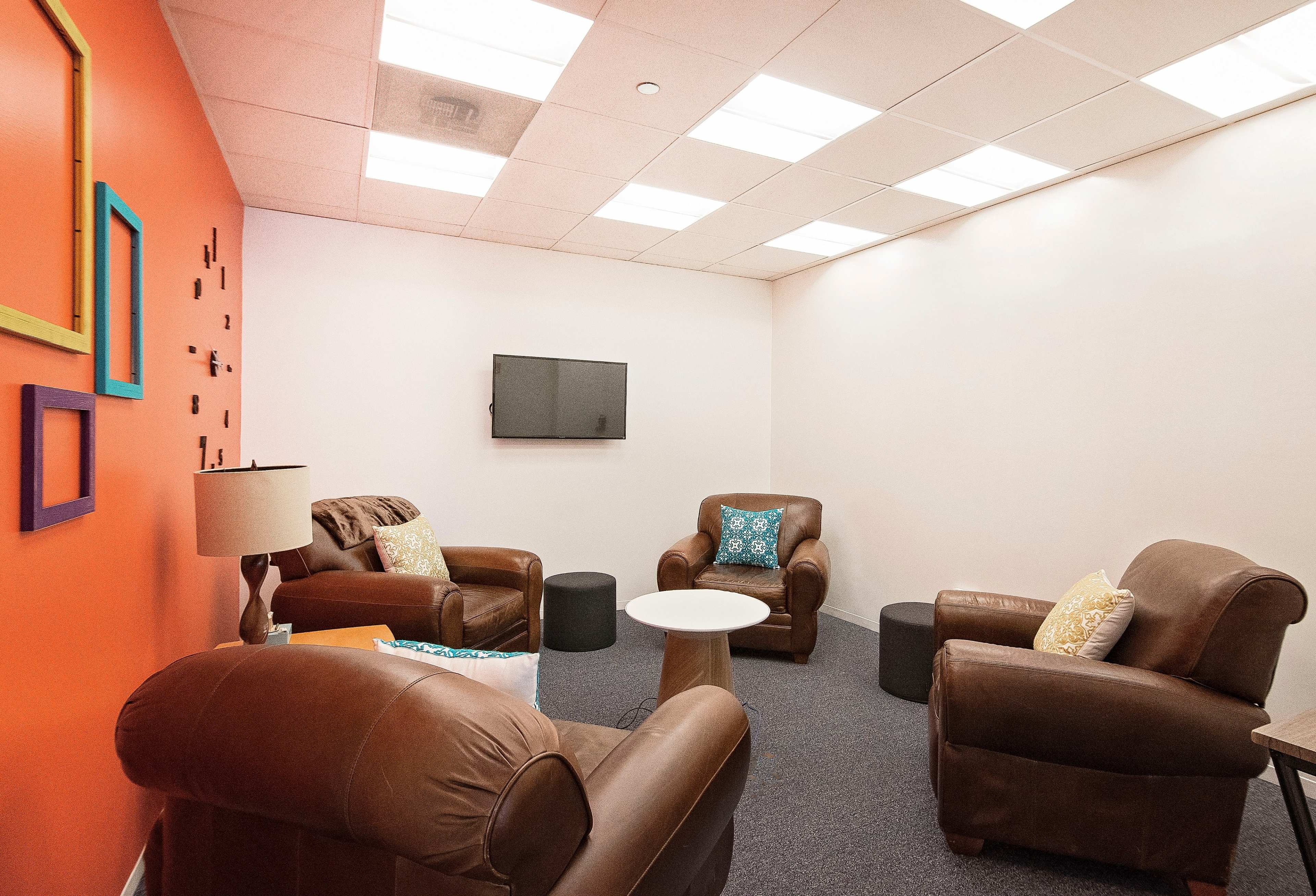 The image shows a small, well-lit lounge area with two brown leather armchairs, a white coffee table, a lamp, and a wall-mounted television.