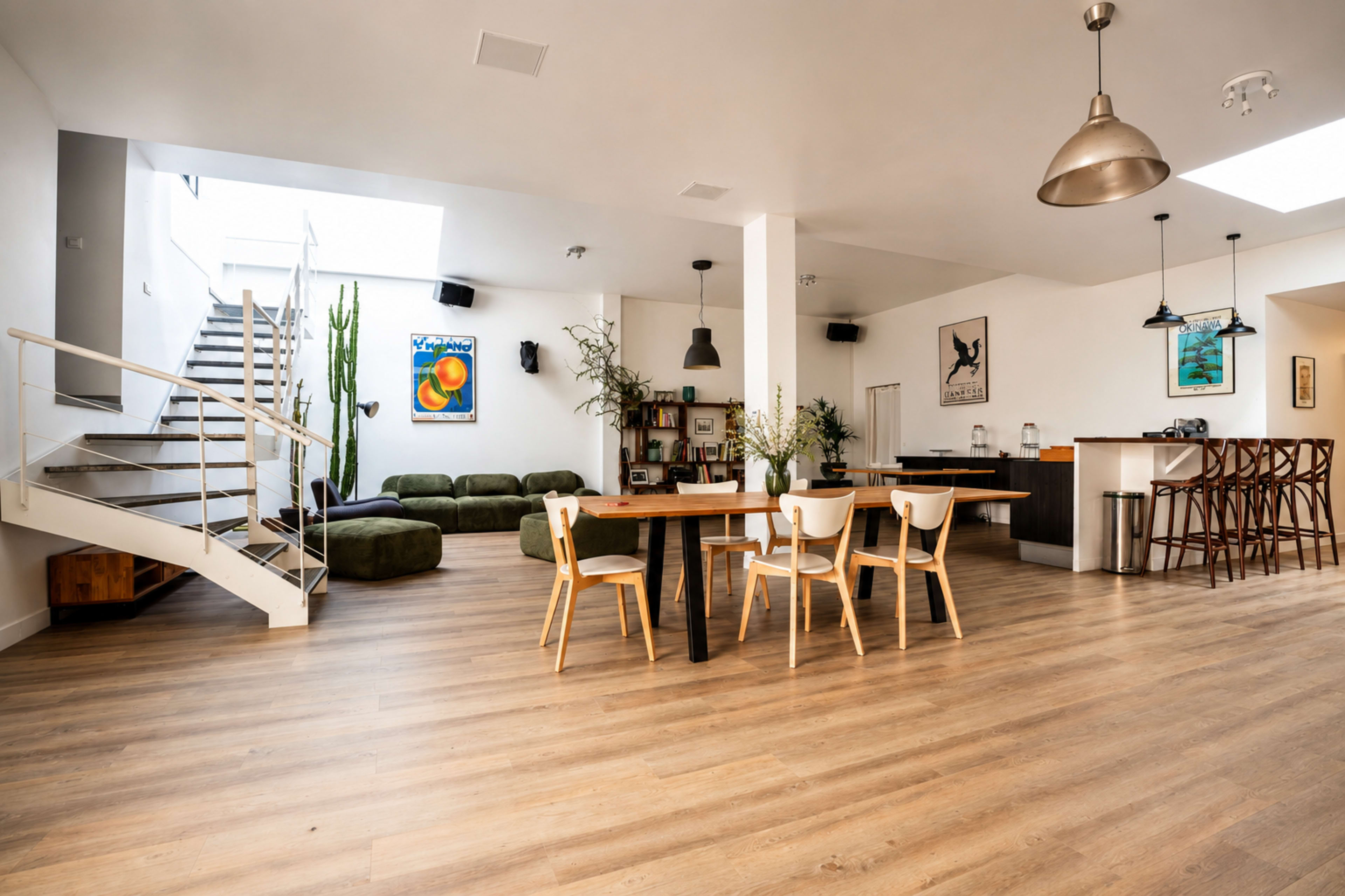 The image shows a spacious, modern interior with a dining table surrounded by chairs, a green sofa, a staircase, and a kitchen area on one side.