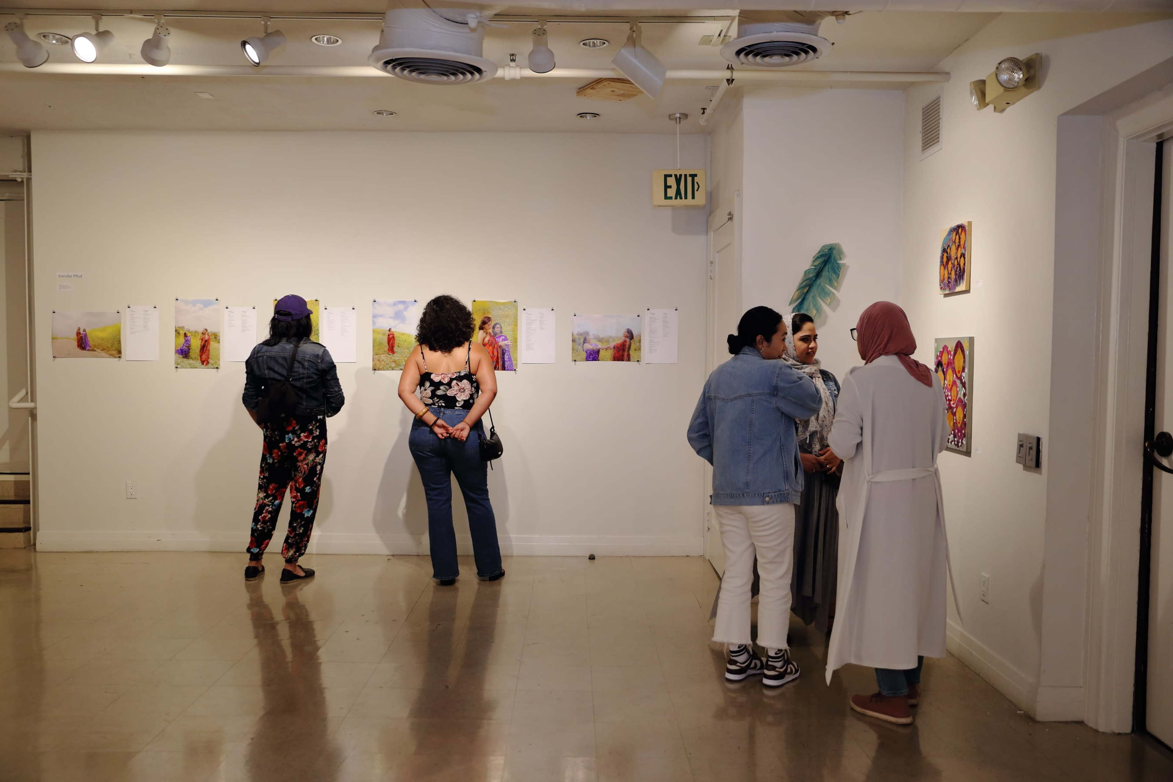 A group of people stands in an art gallery, observing artworks displayed on the wall while others engage in conversation.