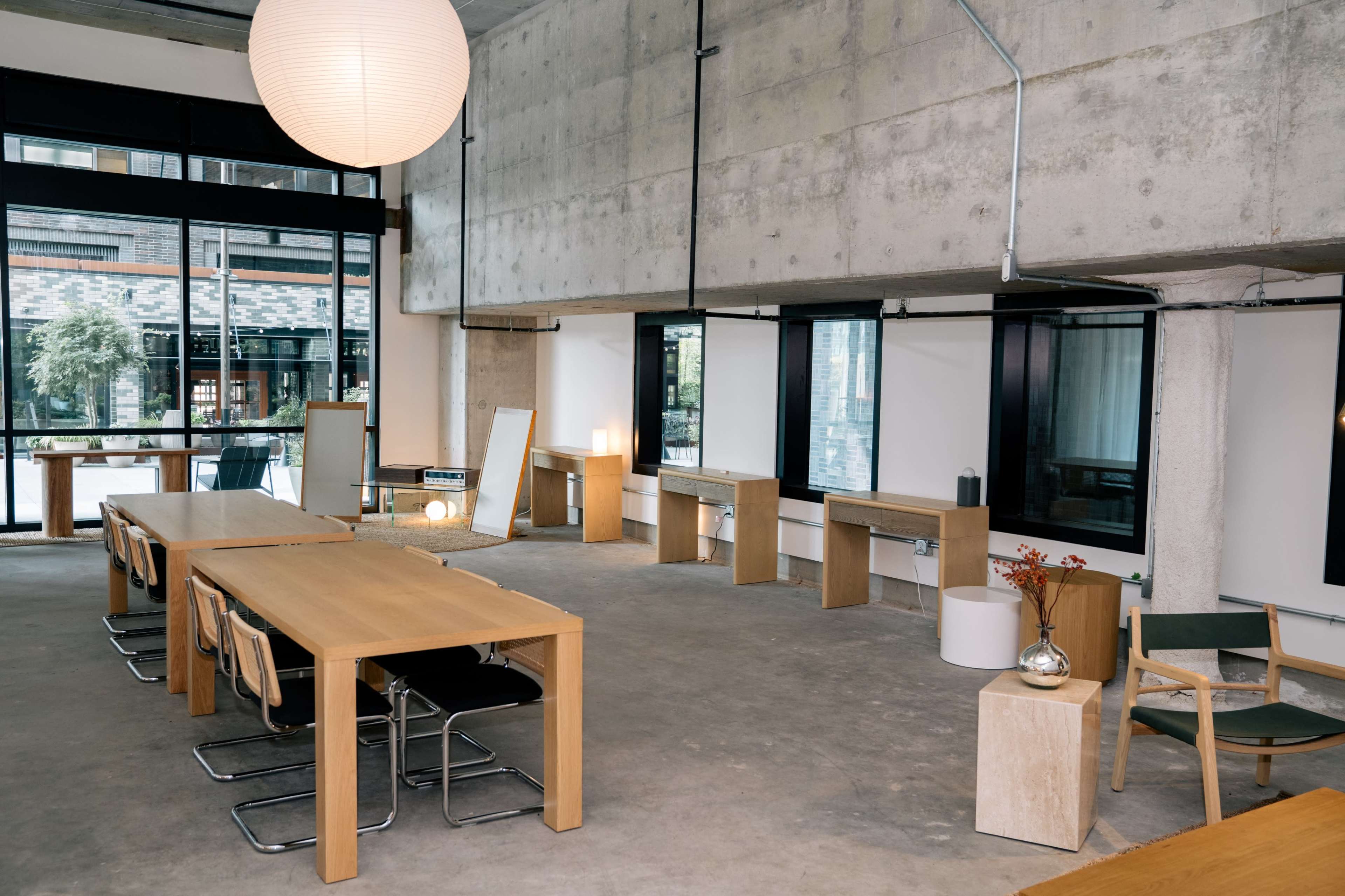 The image shows a modern interior space featuring wooden tables, chairs, and minimalist decor, with large windows allowing natural light to enter.
