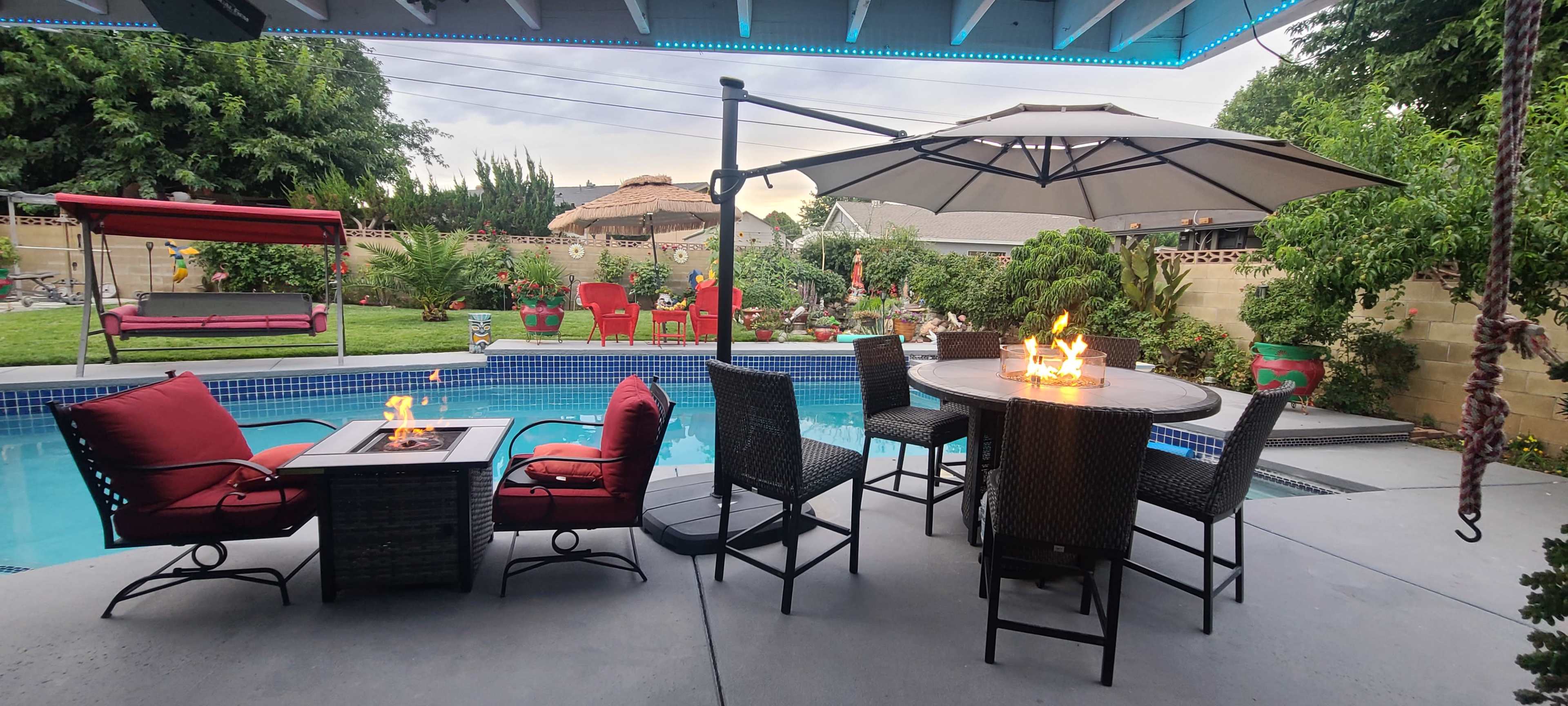 A backyard patio setting with a swimming pool, a large umbrella over a table with flames, and several red chairs and a swing in the garden area.