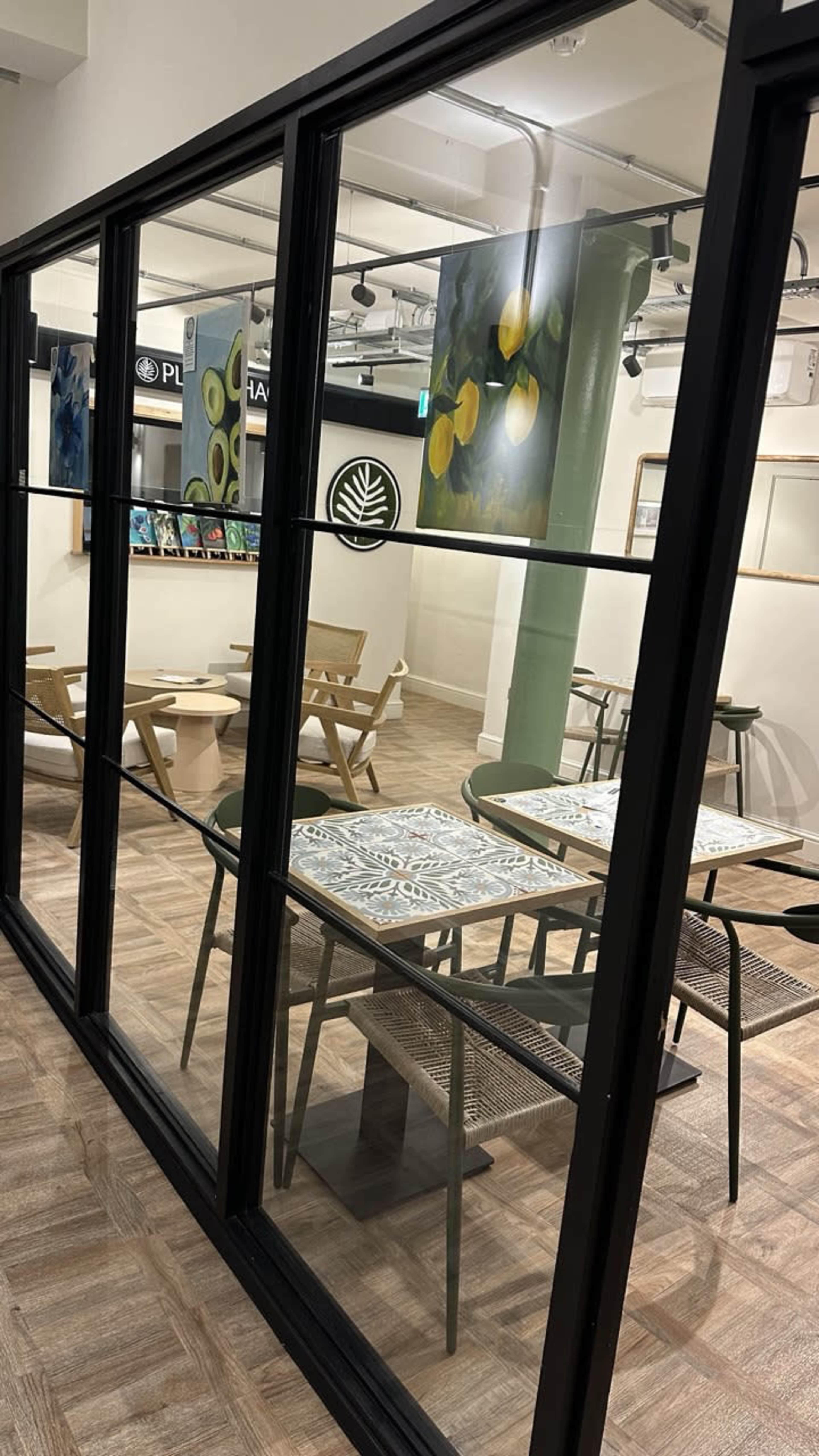 The image shows a spacious indoor café or art studio with tables and chairs arranged behind large glass panels, featuring artwork on the walls.