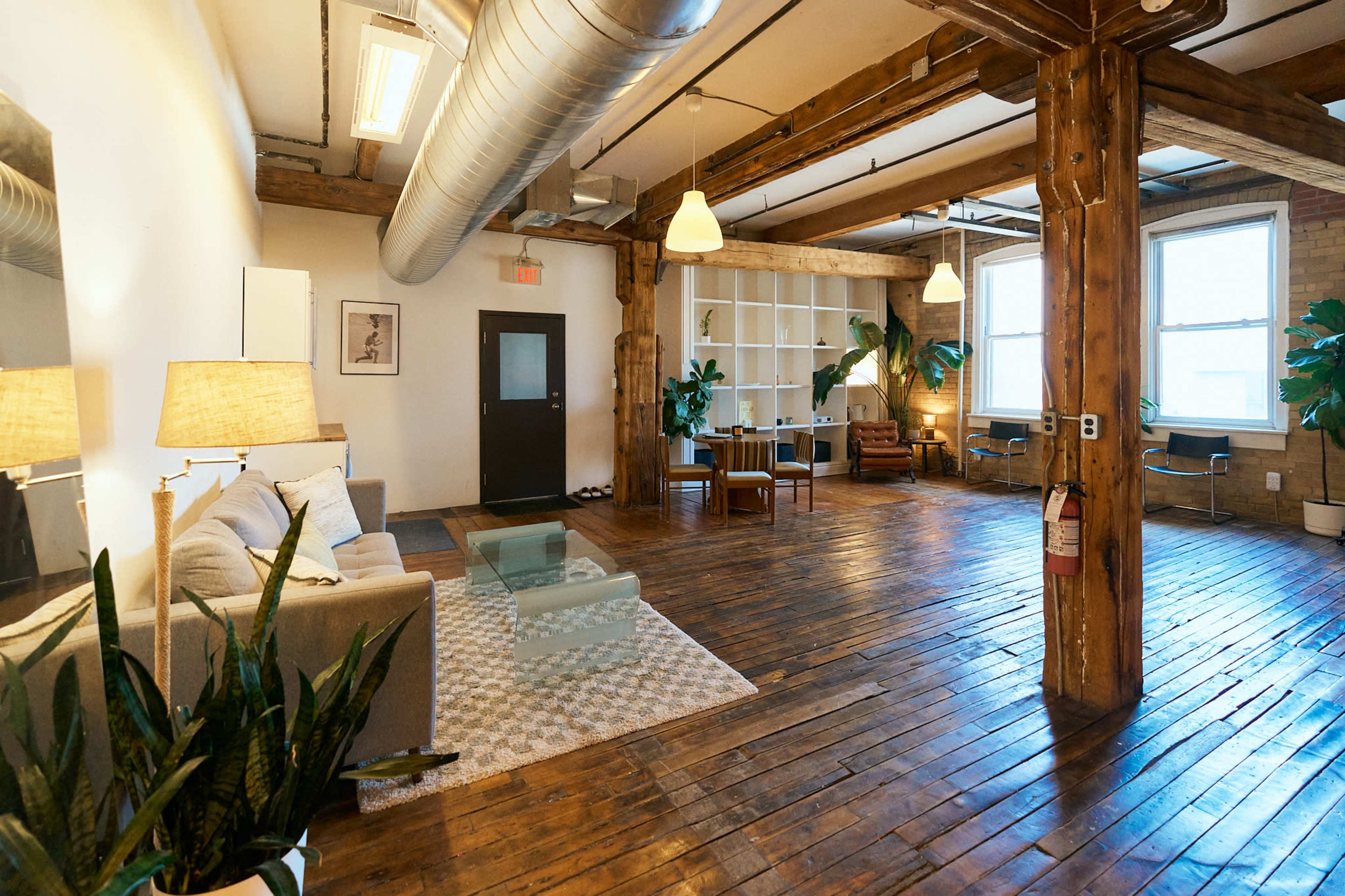 The image shows a spacious, modern living area with wooden beams, a cozy sofa, a coffee table, and plants, featuring large windows and a light, airy atmosphere.