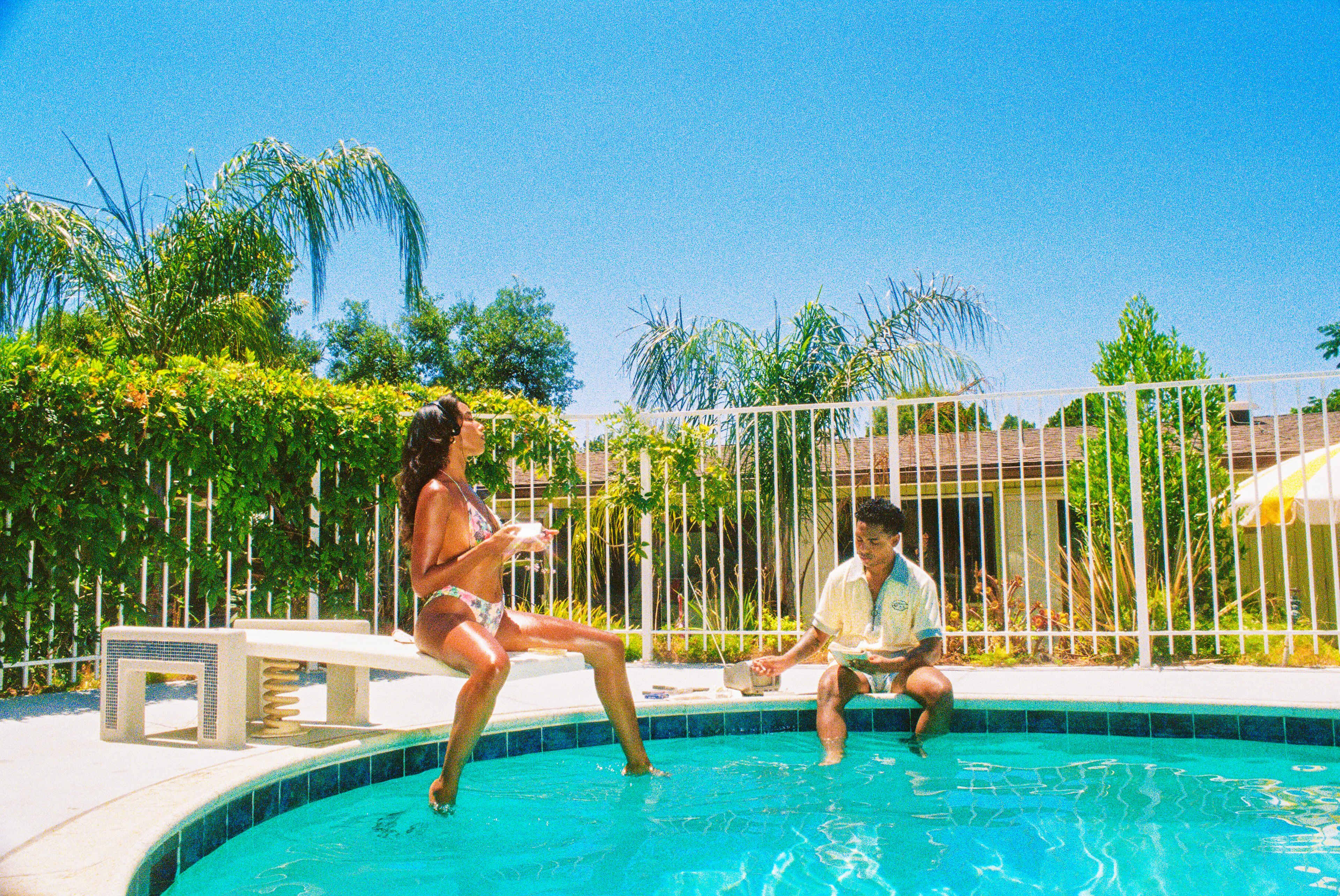 Two people are enjoying a sunny day by a pool, with one sitting on the edge and the other standing in the water.