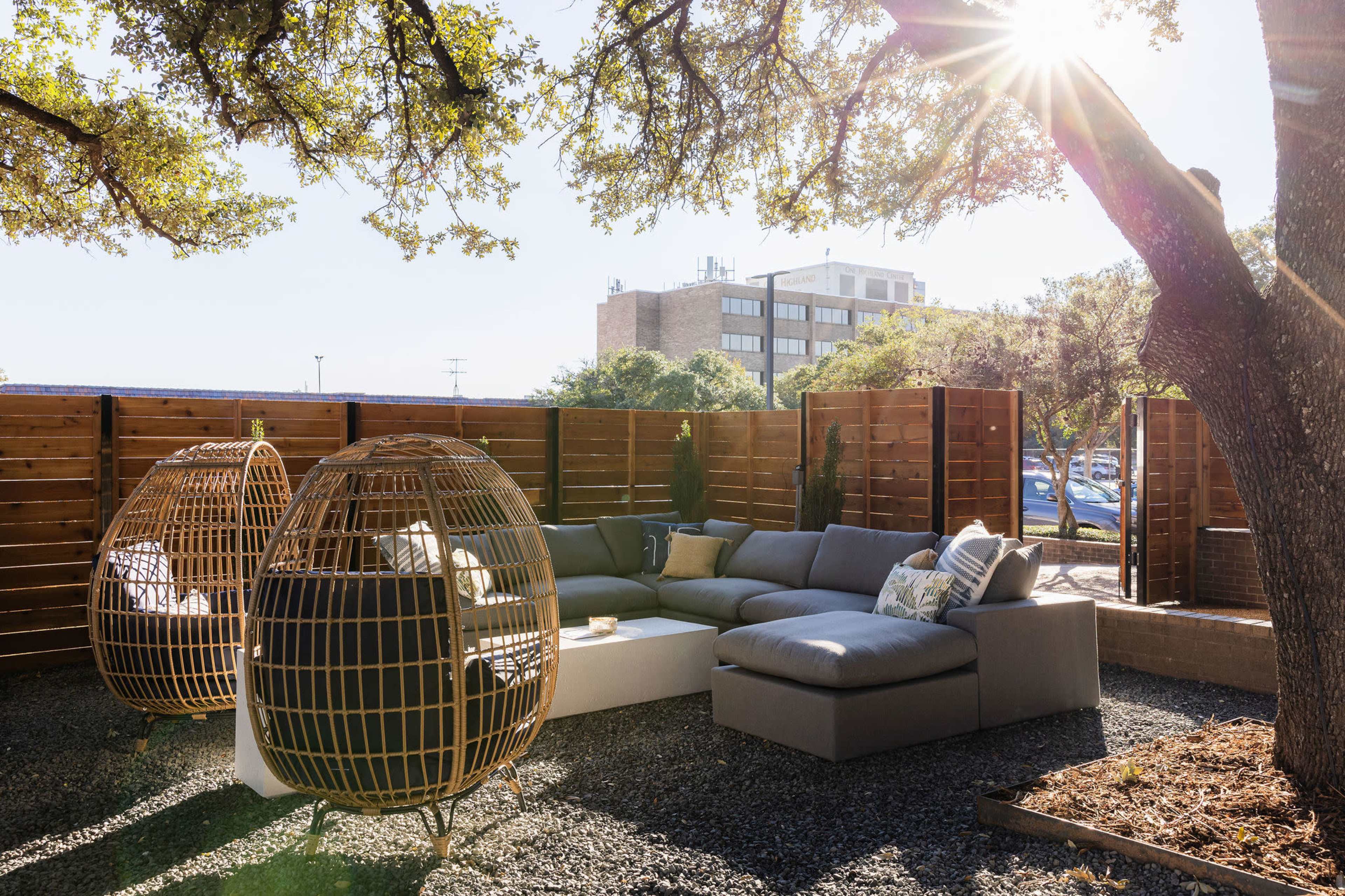 A modern outdoor seating area features a sectional sofa, a coffee table, and two woven lounge chairs surrounded by wooden fences and trees, with sunlight streaming through the leaves.