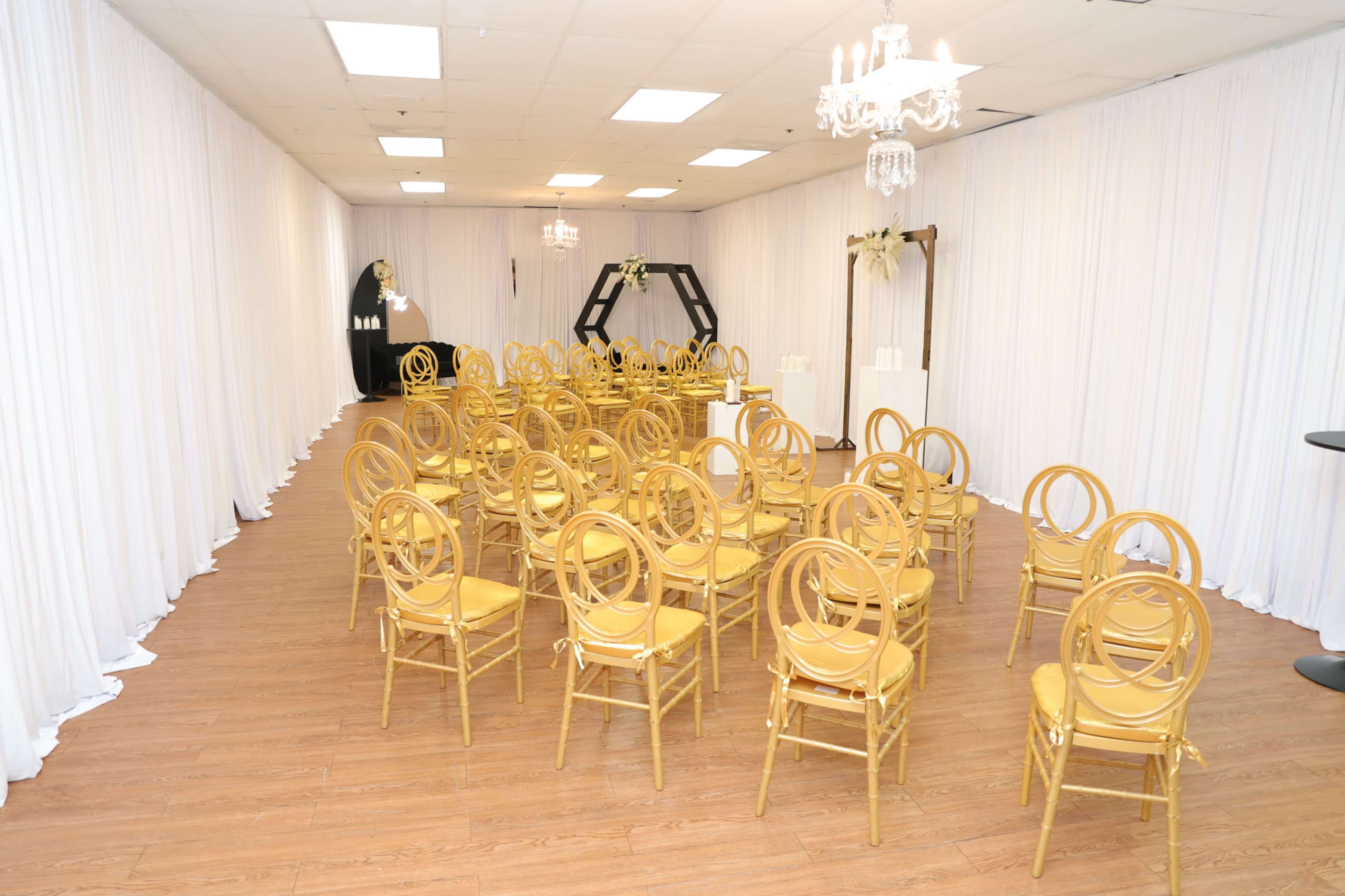 The image shows a spacious room set up with rows of gold chairs facing a decorated backdrop, surrounded by white drapery.
