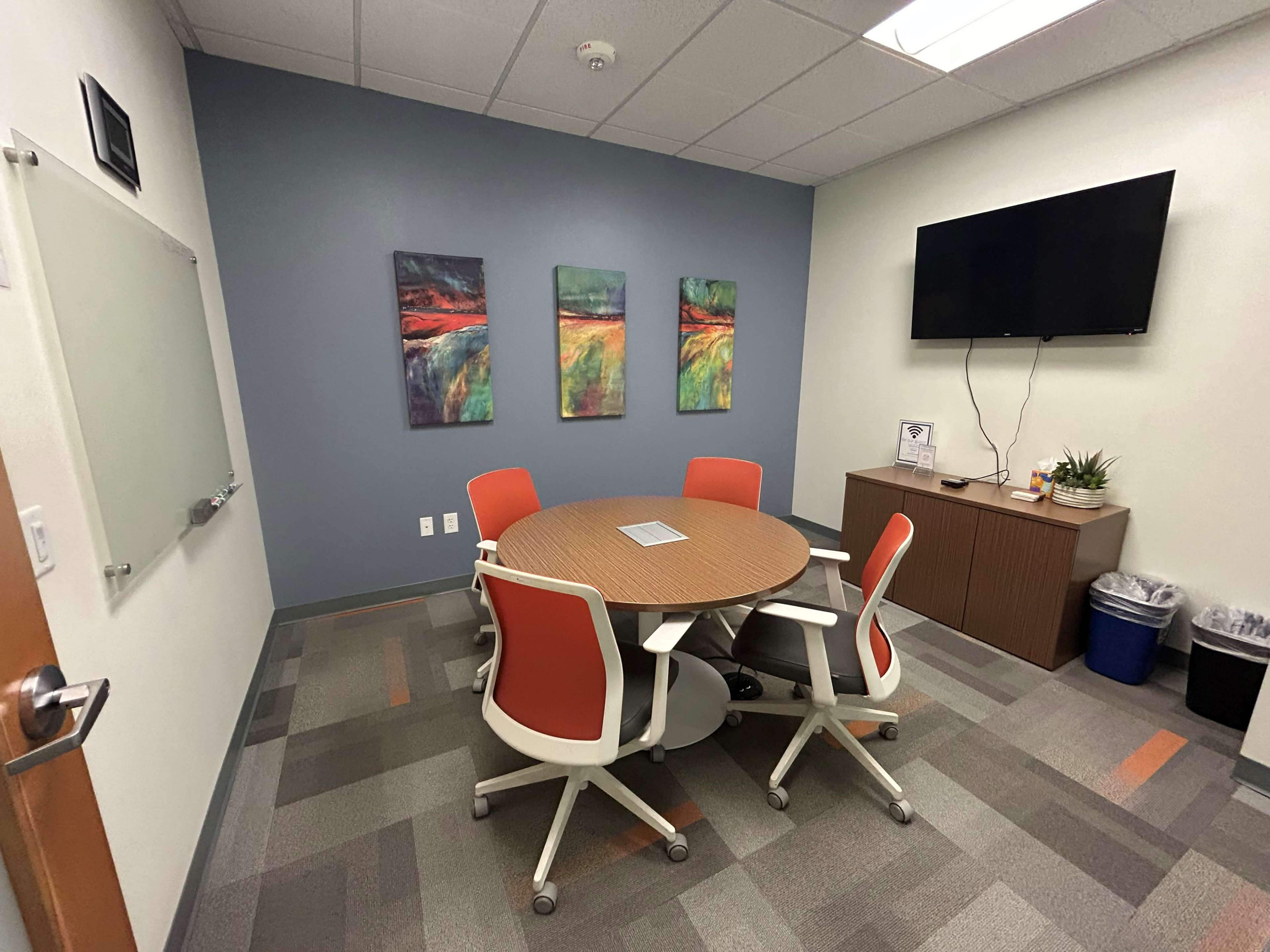 The image shows a small conference room featuring a round table with five chairs, three abstract paintings on the wall, a television mounted in one corner, and a cabinet with a plant and waste bin.