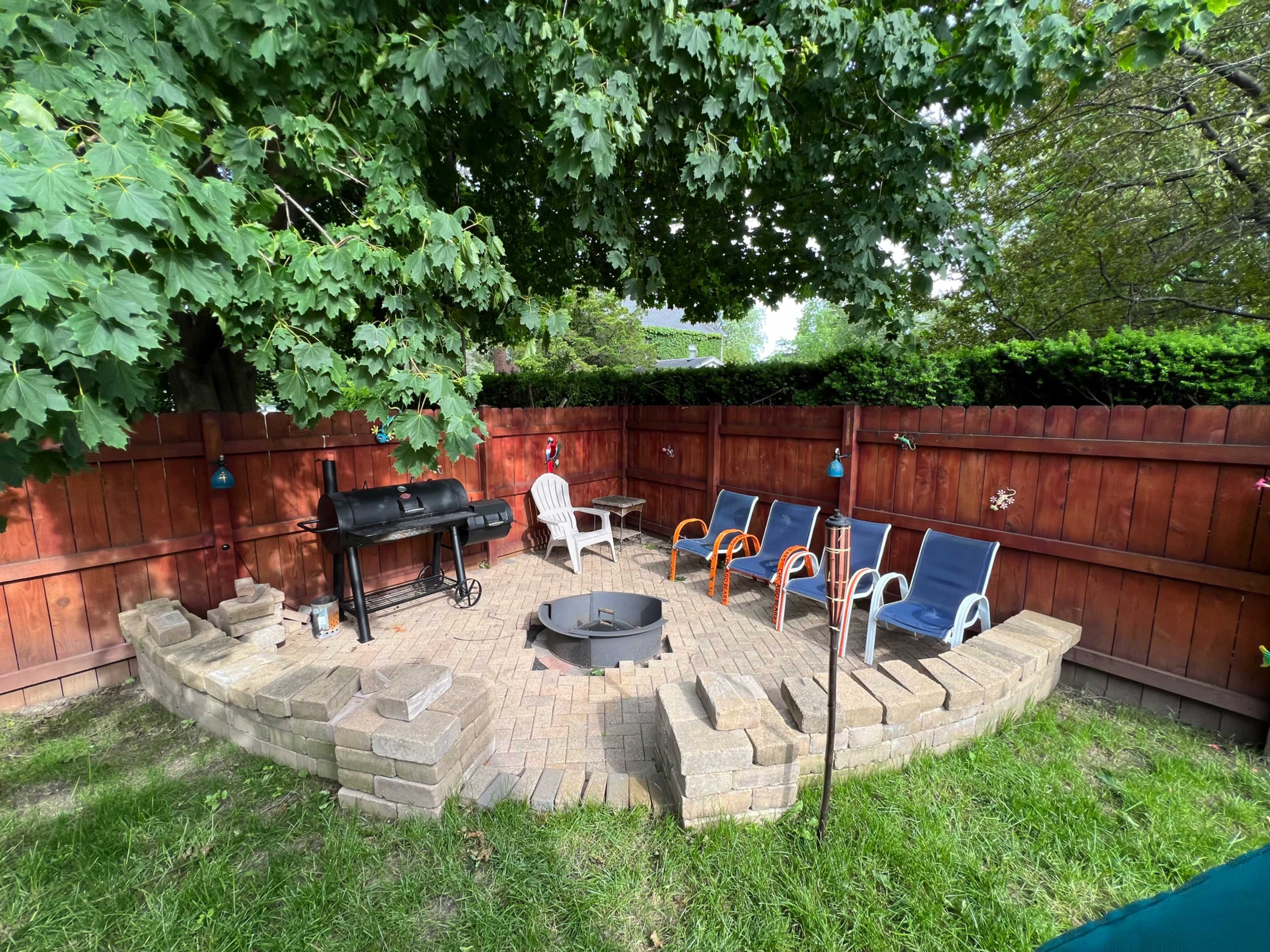 The image shows a backyard patio area with a fire pit surrounded by stone seating, a charcoal grill, and colorful chairs, enclosed by a wooden fence and shaded by trees.