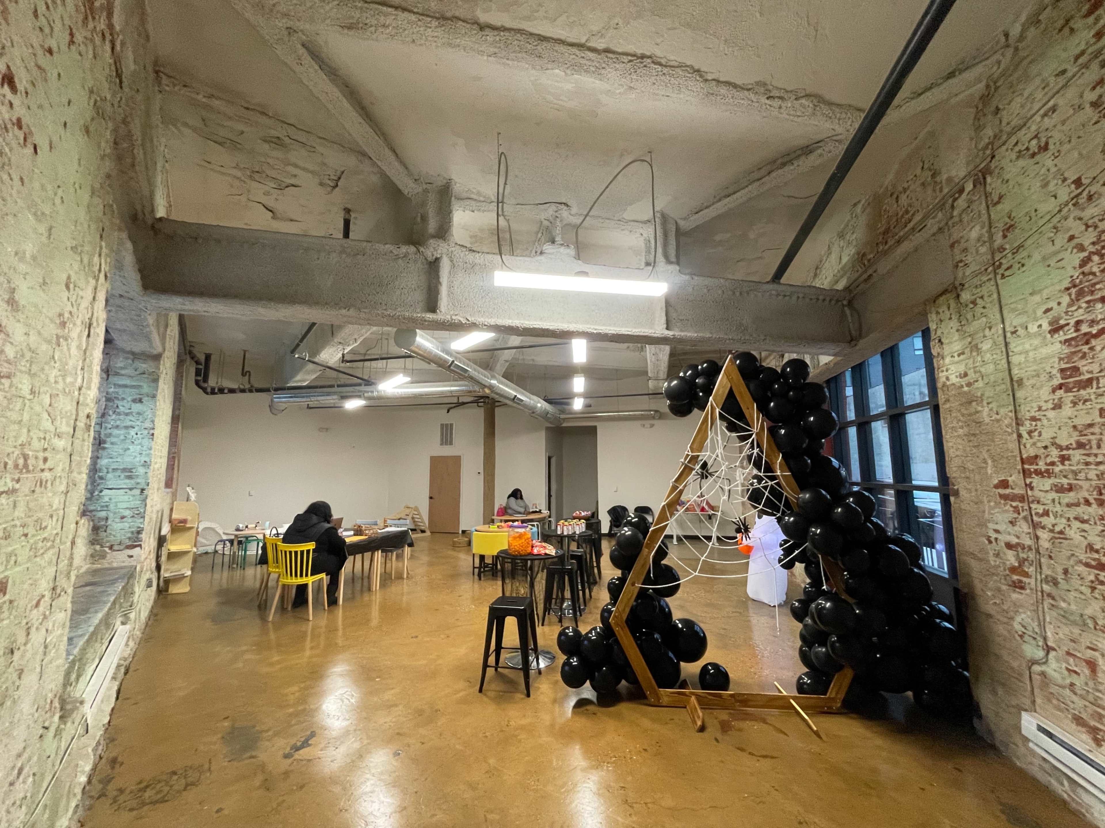 An industrial-style room with exposed brick walls, a large window, and a black balloon arch decorated with spider web, while several people are engaged in activities at tables.