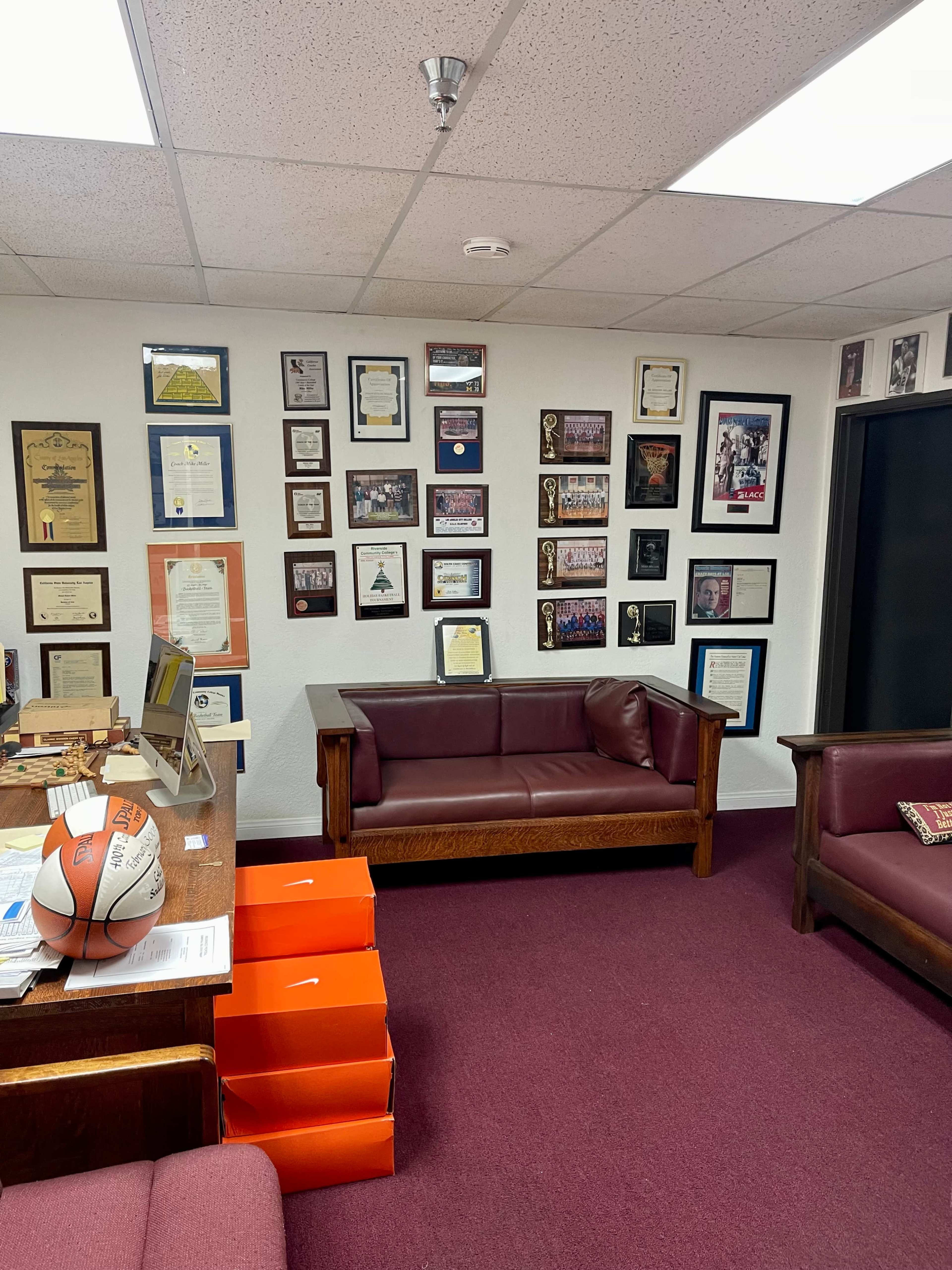 A room with a dark red carpet, a desk covered in papers, two brown couches, and walls adorned with various certificates and framed photographs related to sports achievements.