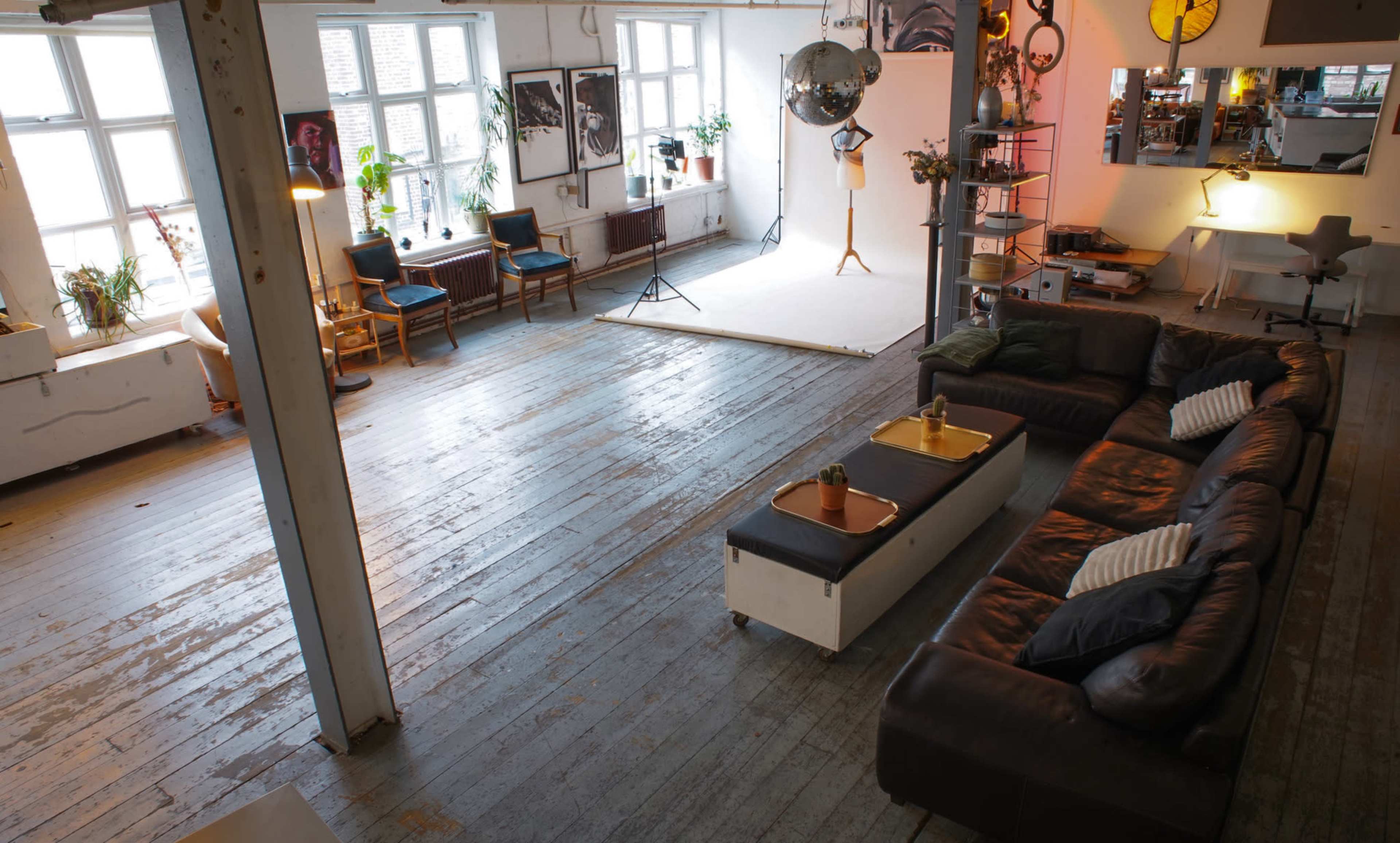 The image shows a spacious interior with wooden floors, large windows, a sectional sofa, and various studio props arranged for photography.