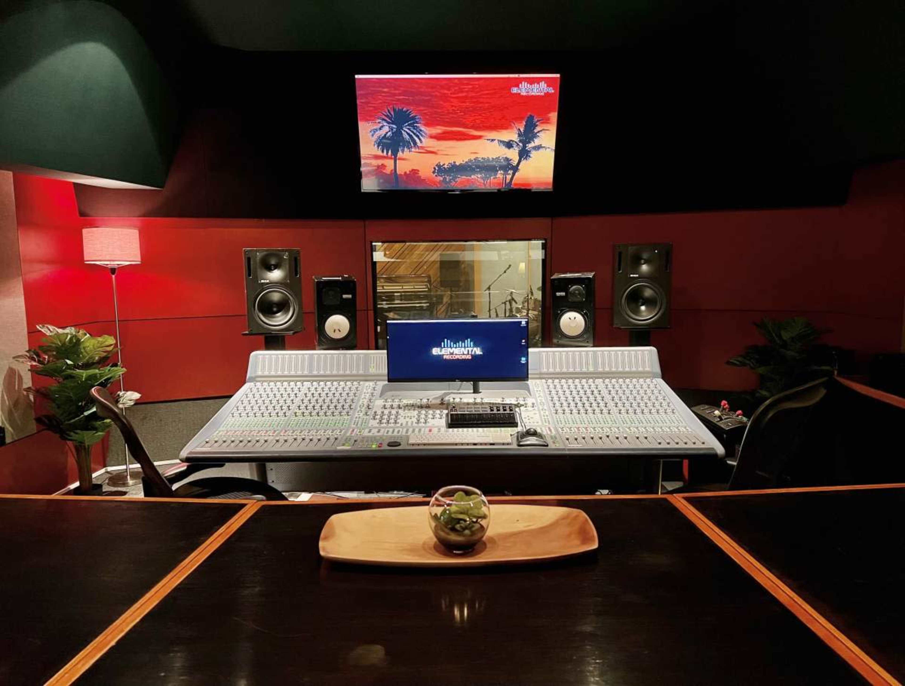 A recording studio is shown with a large mixing console, two speakers on each side, and a digital screen displaying an image in the background.