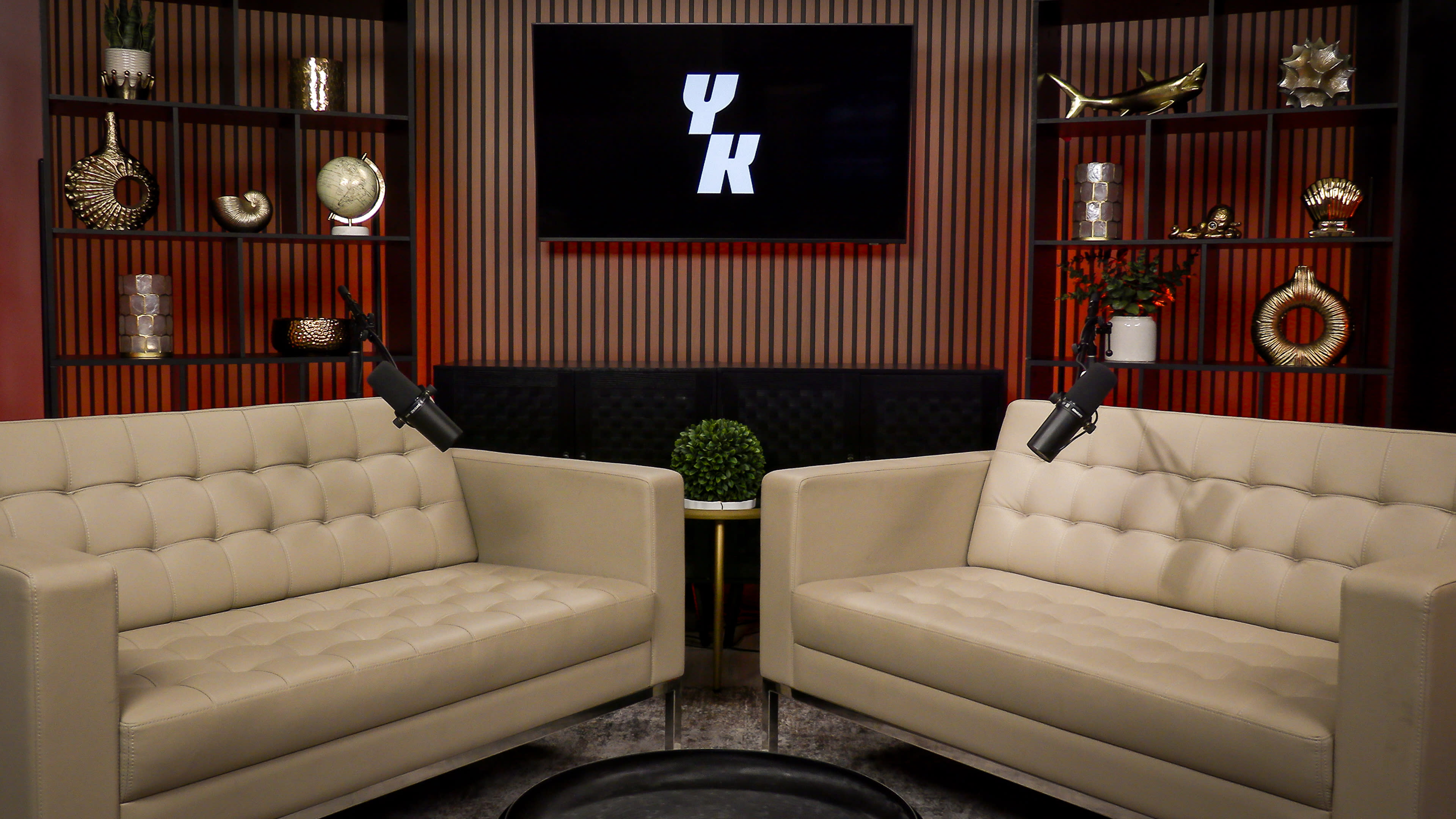 The image shows a modern studio setup featuring two beige sofas, a small table, and a backdrop with decorative shelves and a screen displaying "YK."