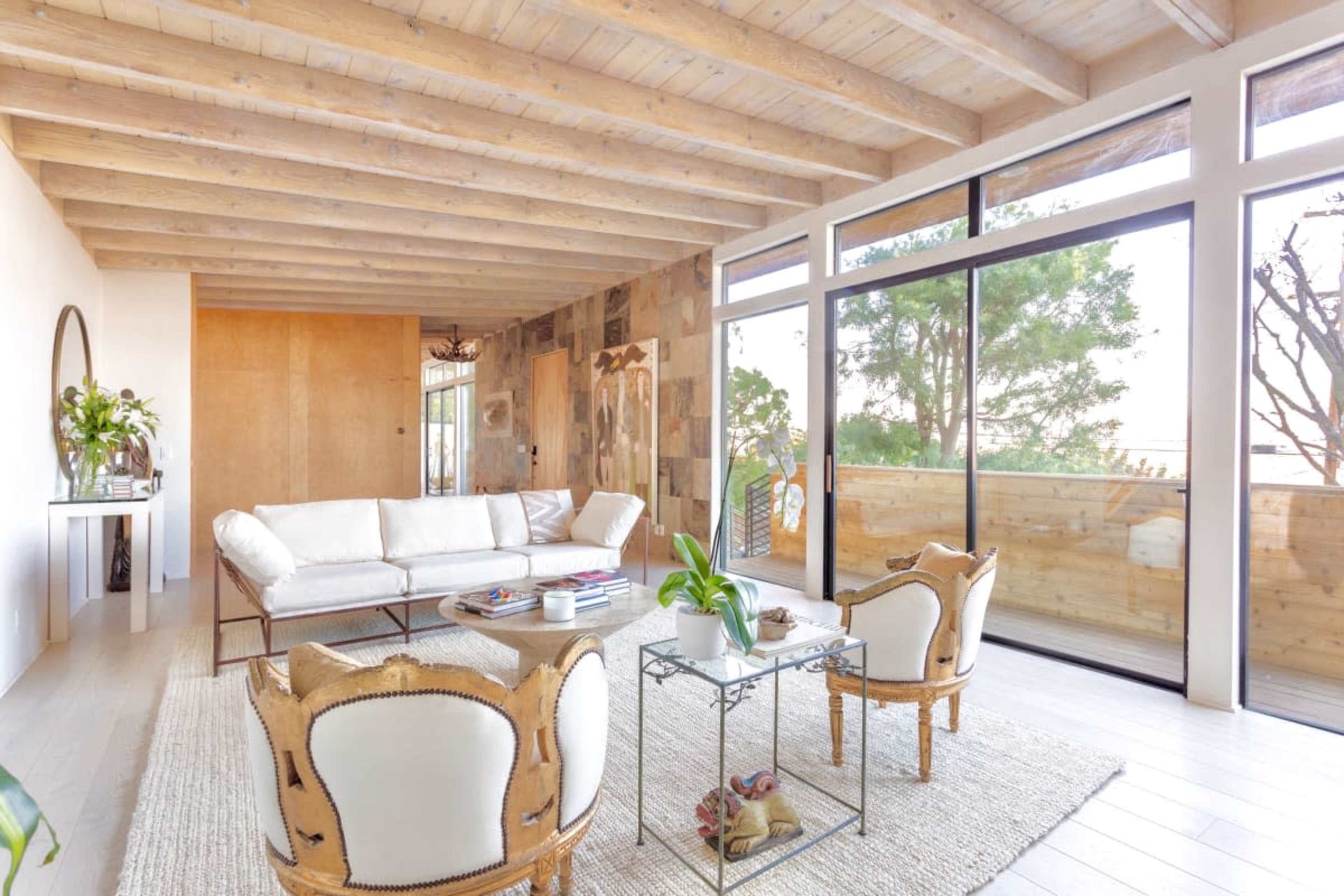 The image shows a modern living room with a white sofa, two patterned chairs, a coffee table, and large windows facing a wooden deck and outdoor greenery.