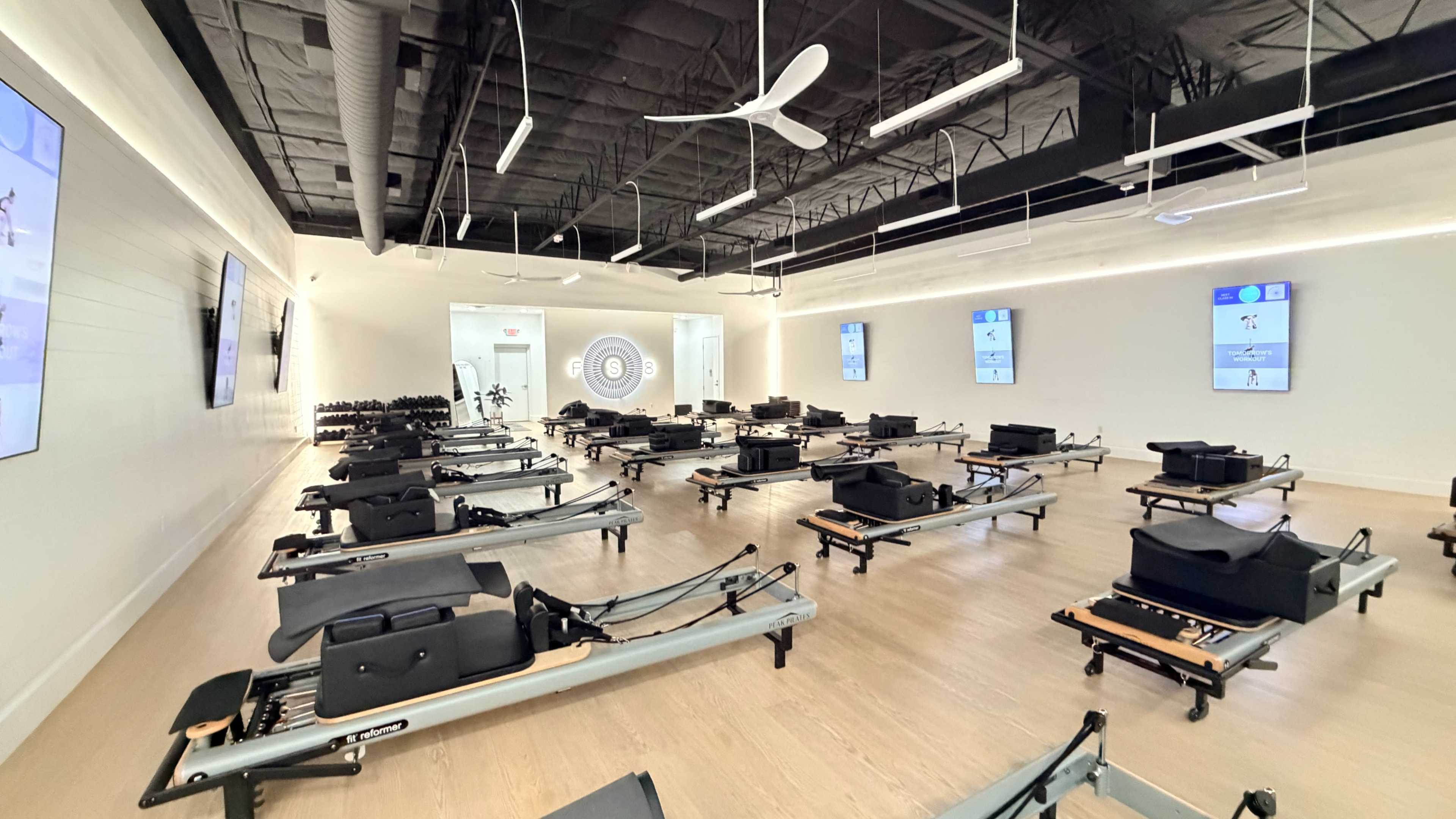 The image shows a spacious fitness studio equipped with several pilates reformers arranged in rows and large screens displaying workout instructions.