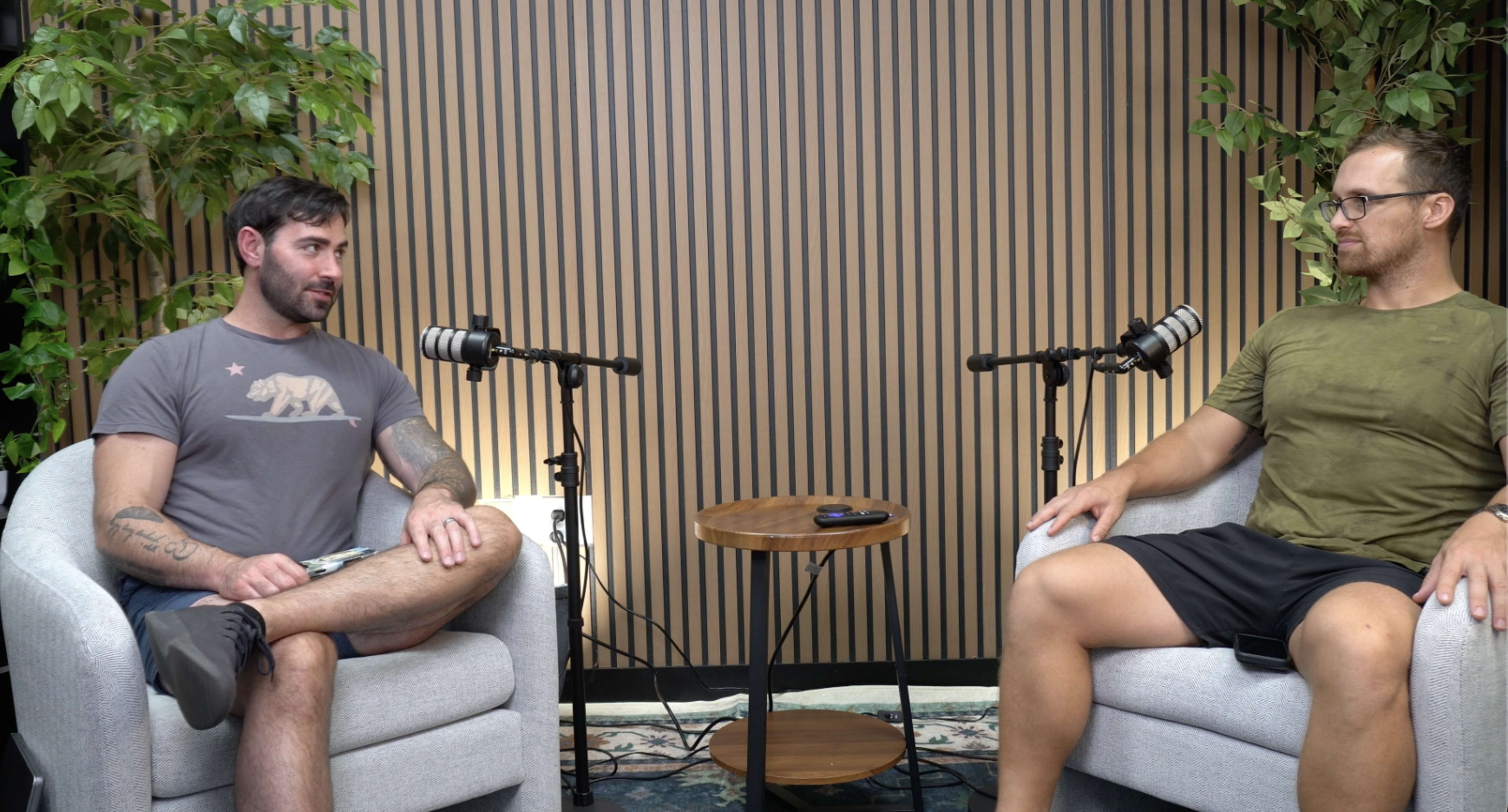Two men sit in light-colored armchairs across from each other in a podcast studio with microphones, a small wooden table, and greenery in the background.