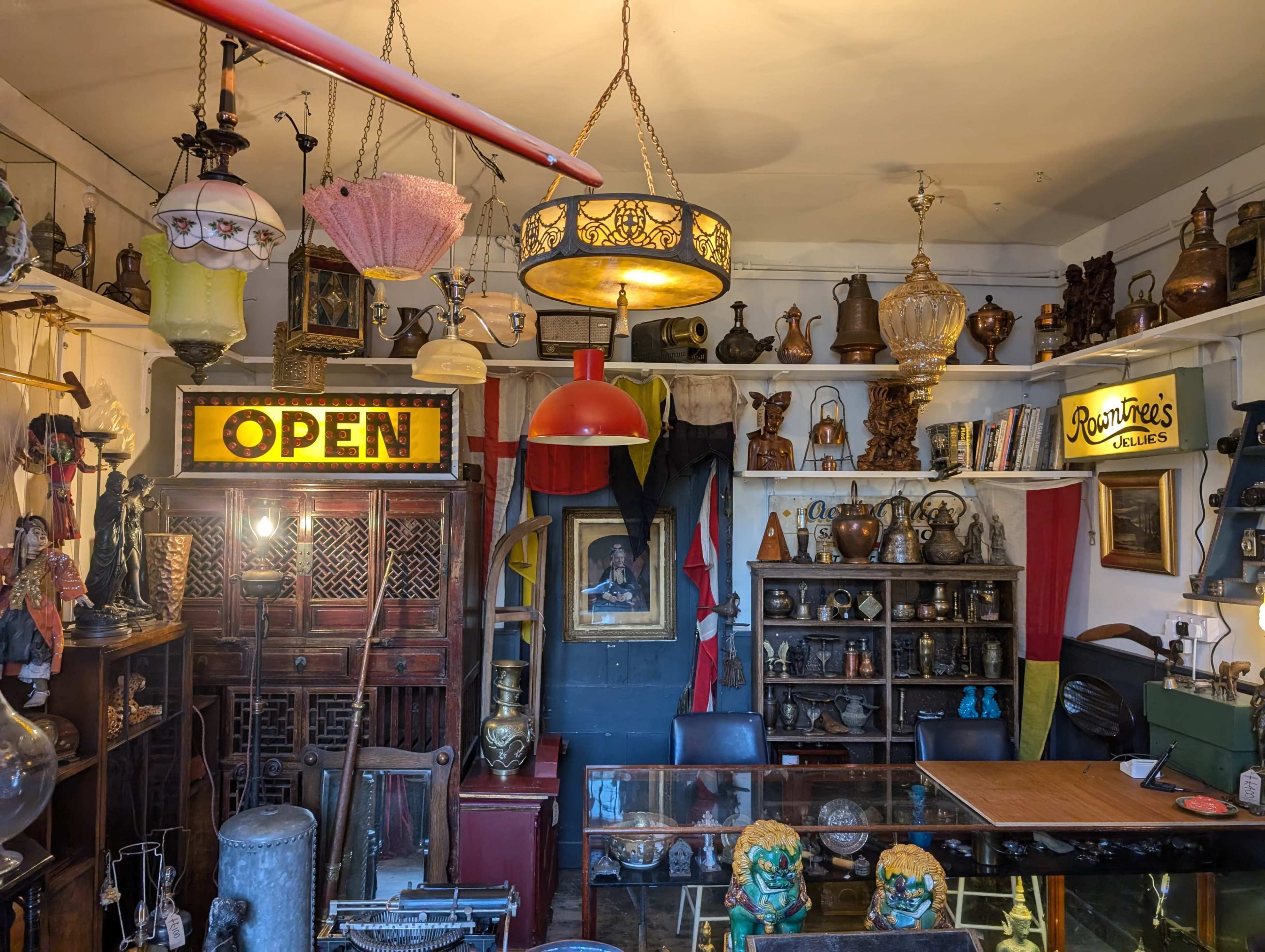 A small antique shop features various vintage items, including lamps, sculptures, and framed art, with an illuminated "OPEN" sign displayed prominently.