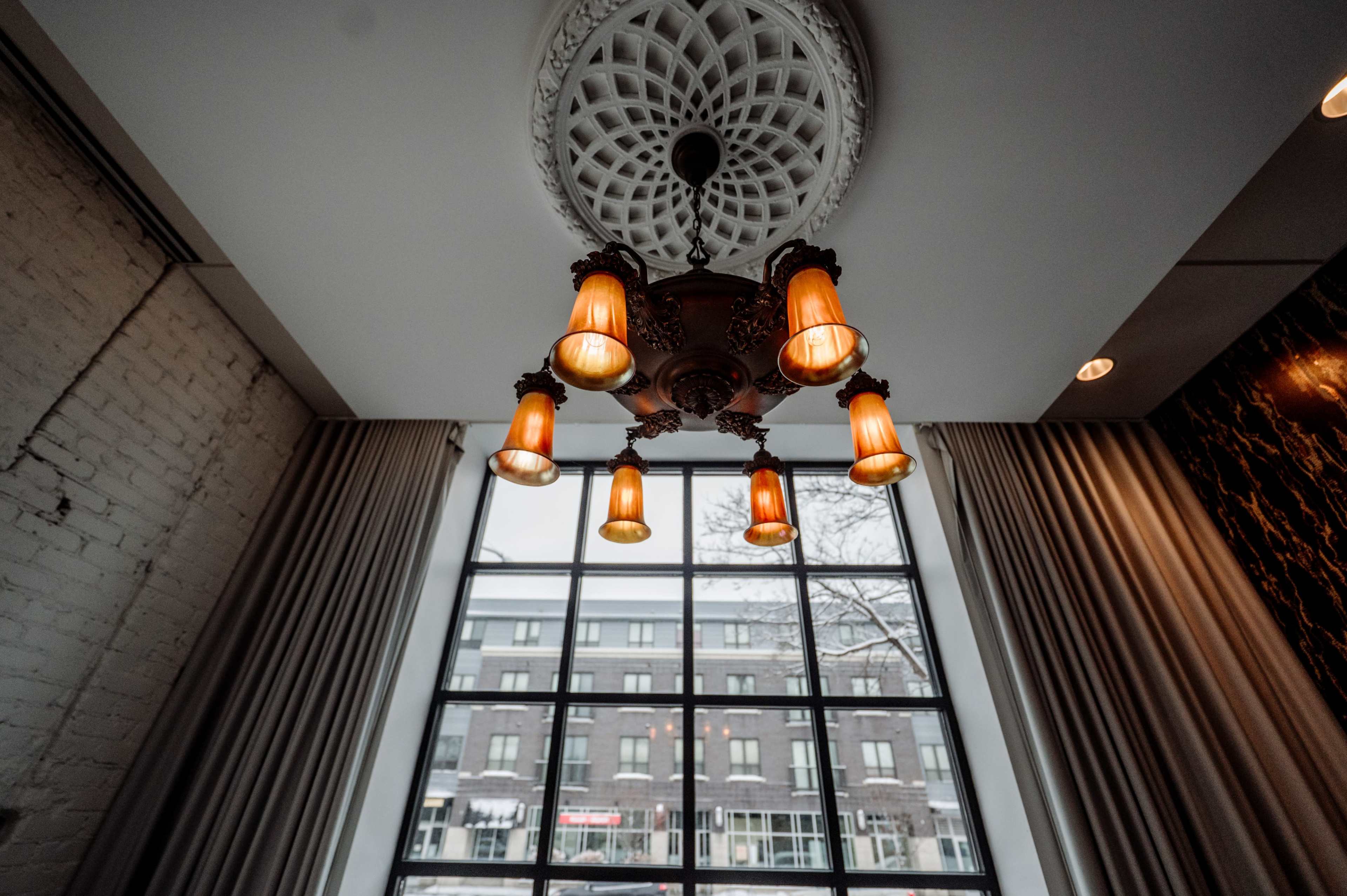 The image shows a decorative ceiling light fixture with multiple amber-colored bulbs hanging from an ornate ceiling medallion, illuminated against a backdrop of large windows and curtains.