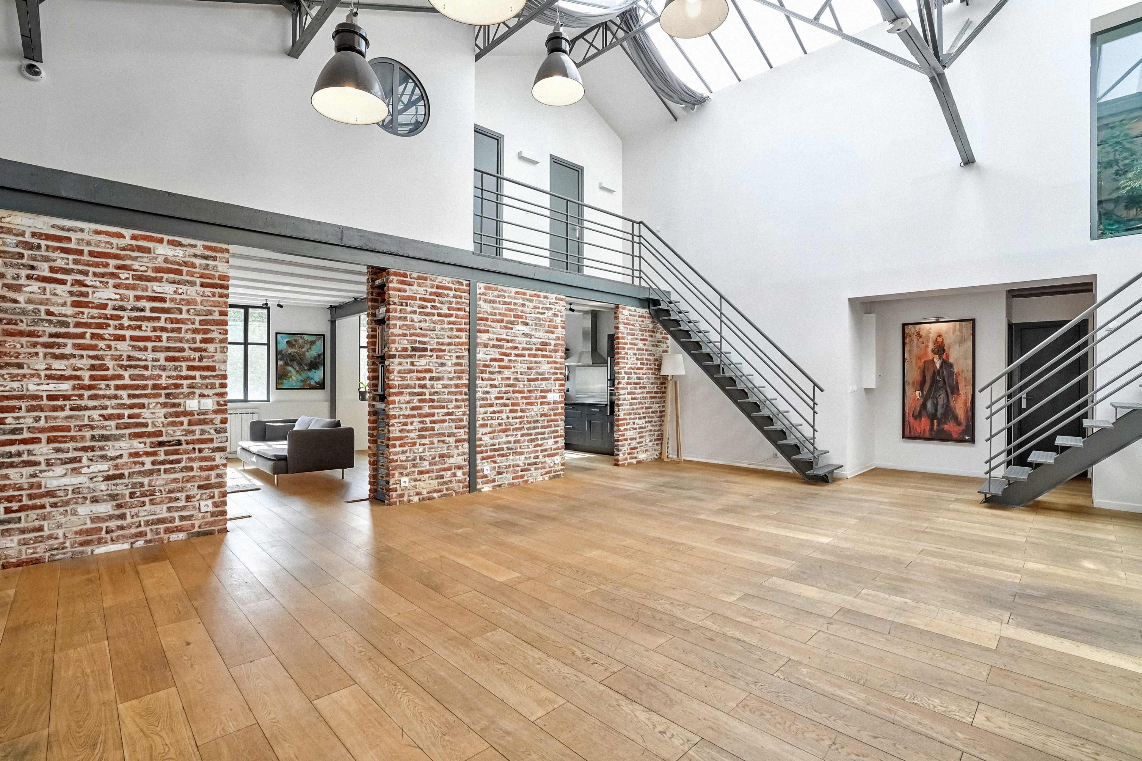 The image shows a spacious, modern interior featuring a double-height ceiling, exposed brick walls, and a wooden floor with stairs leading to an upper level.