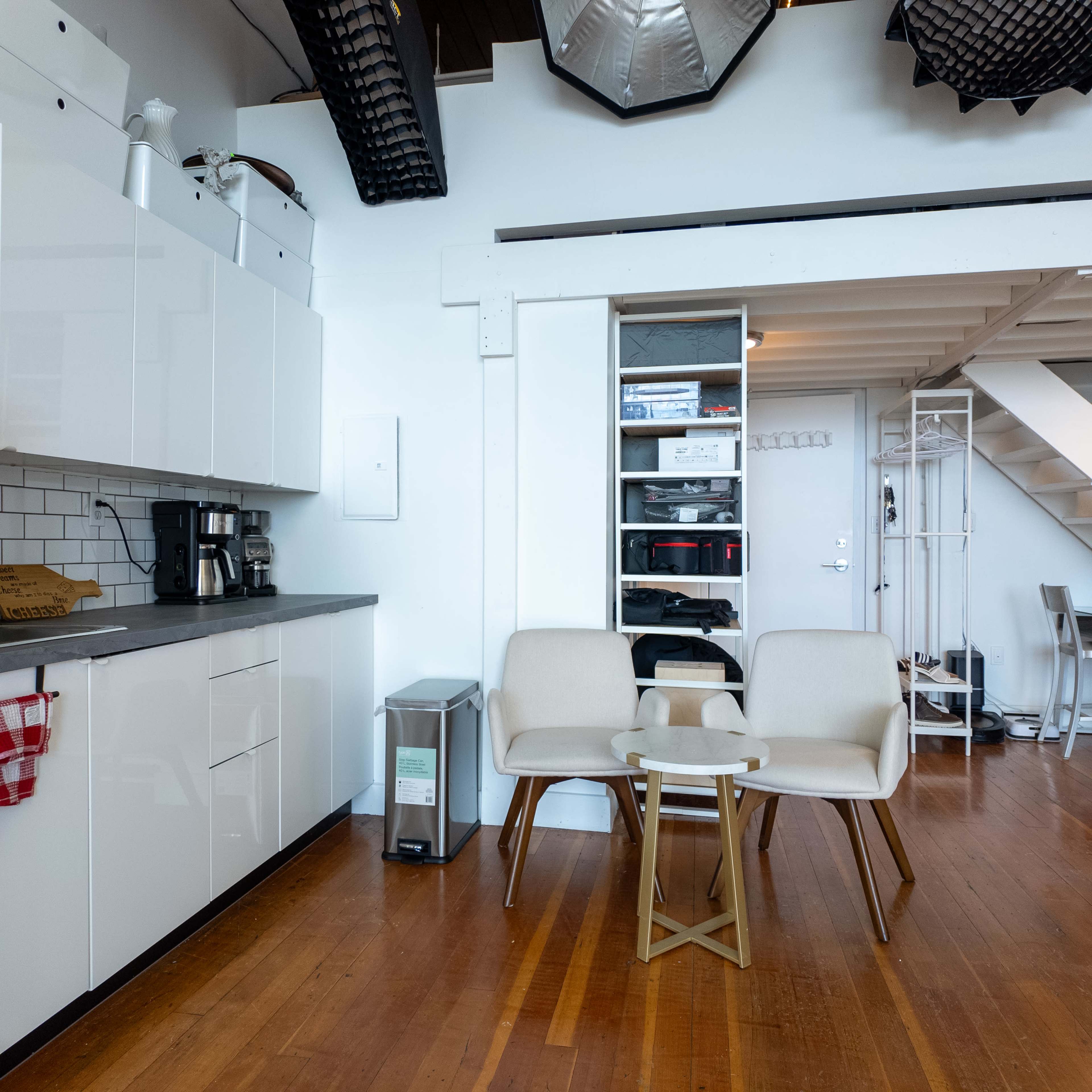 A bright kitchen and living area with white cabinets, a coffee maker, two cream-colored chairs, a small table, and shelving with various items.