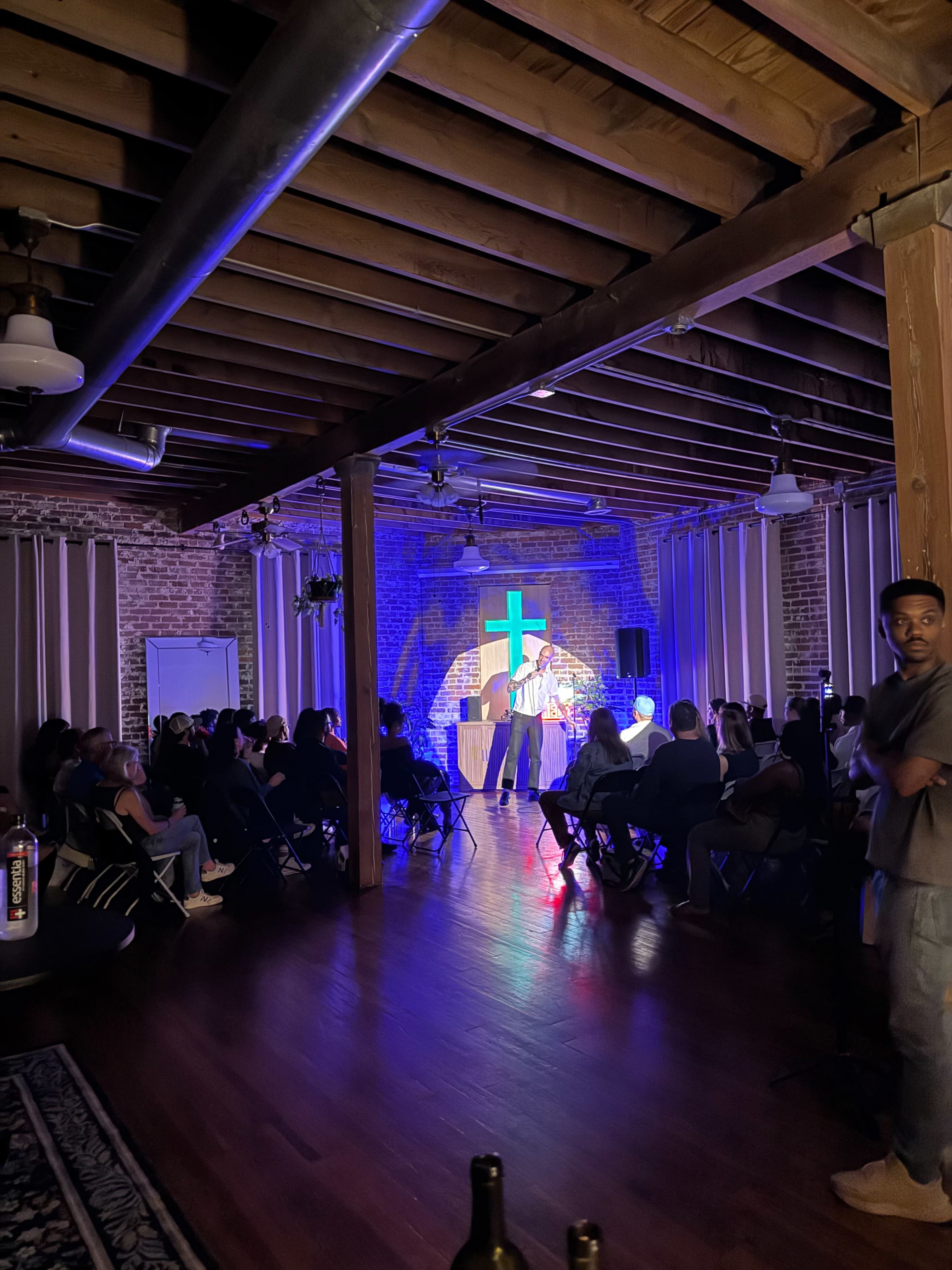 A performer stands on stage in front of a crowd in a dimly lit venue decorated with exposed brick and wooden beams.