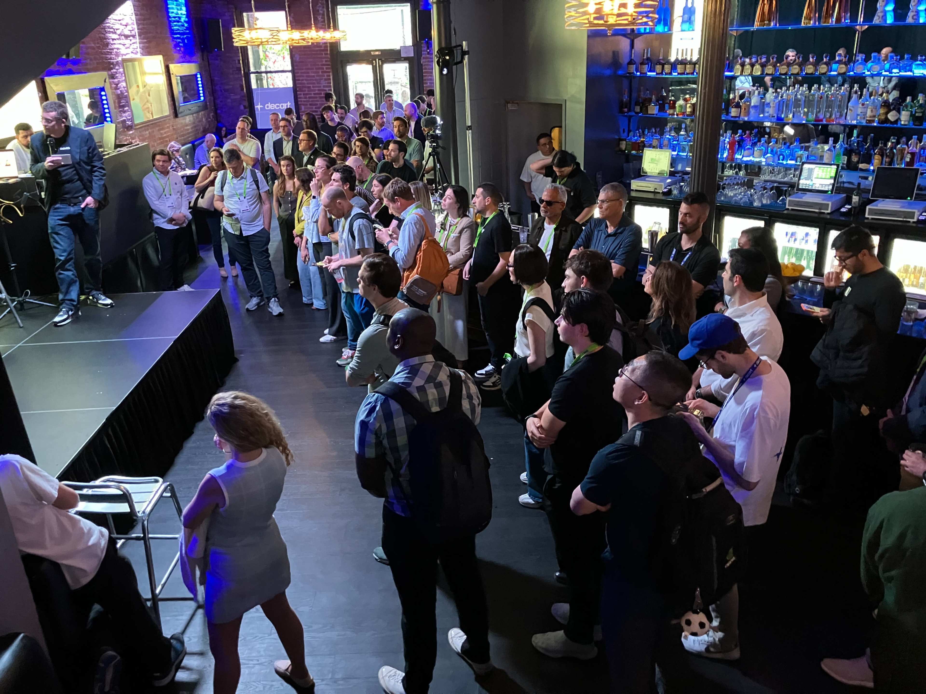 A crowd of people stands in a dimly lit venue, facing a speaker on an elevated platform, while bar shelves filled with bottles are visible in the background.