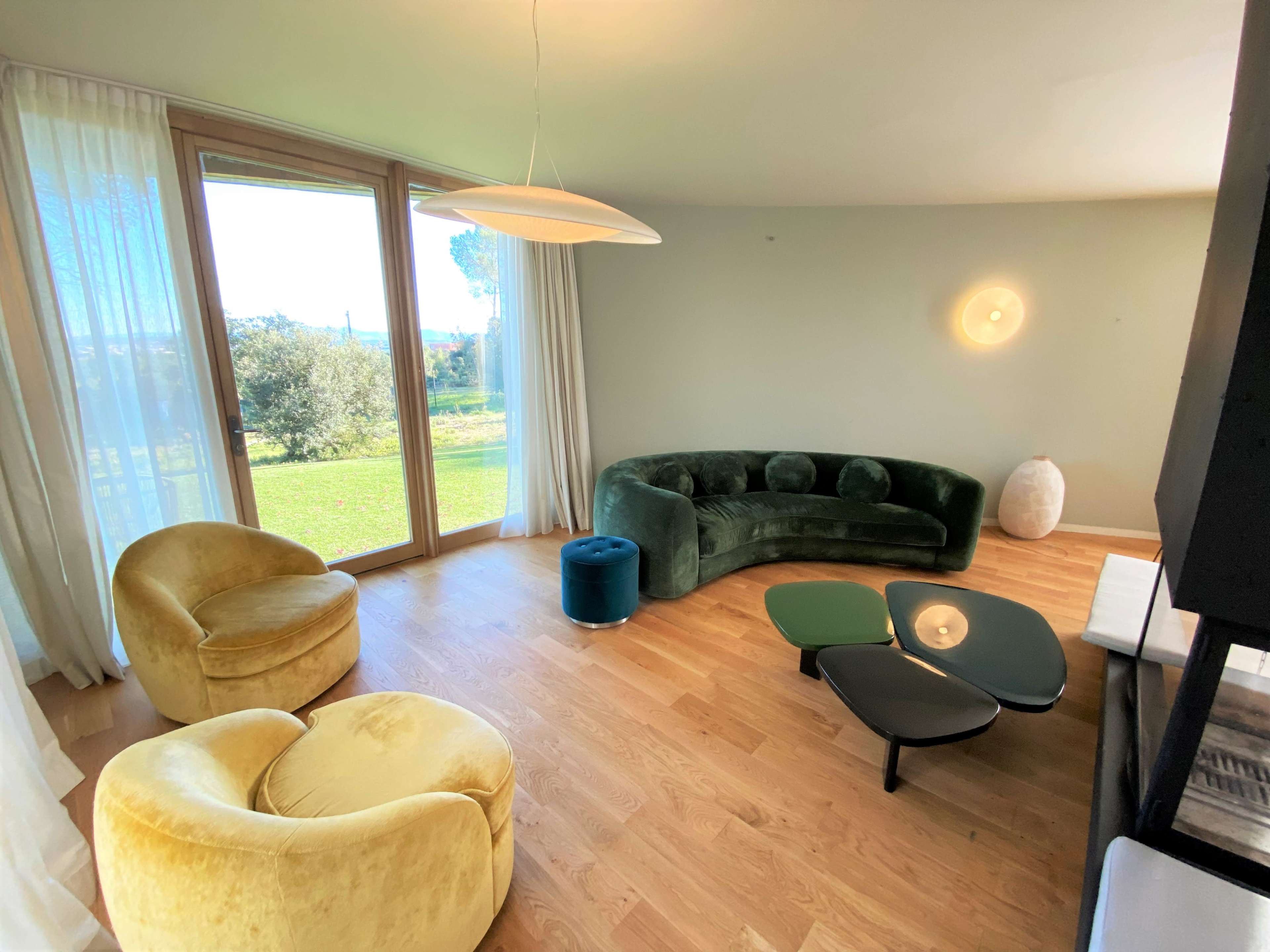 The interior features a modern living room with a green curved sofa, two yellow armchairs, a small blue ottoman, and a coffee table set on a wooden floor.