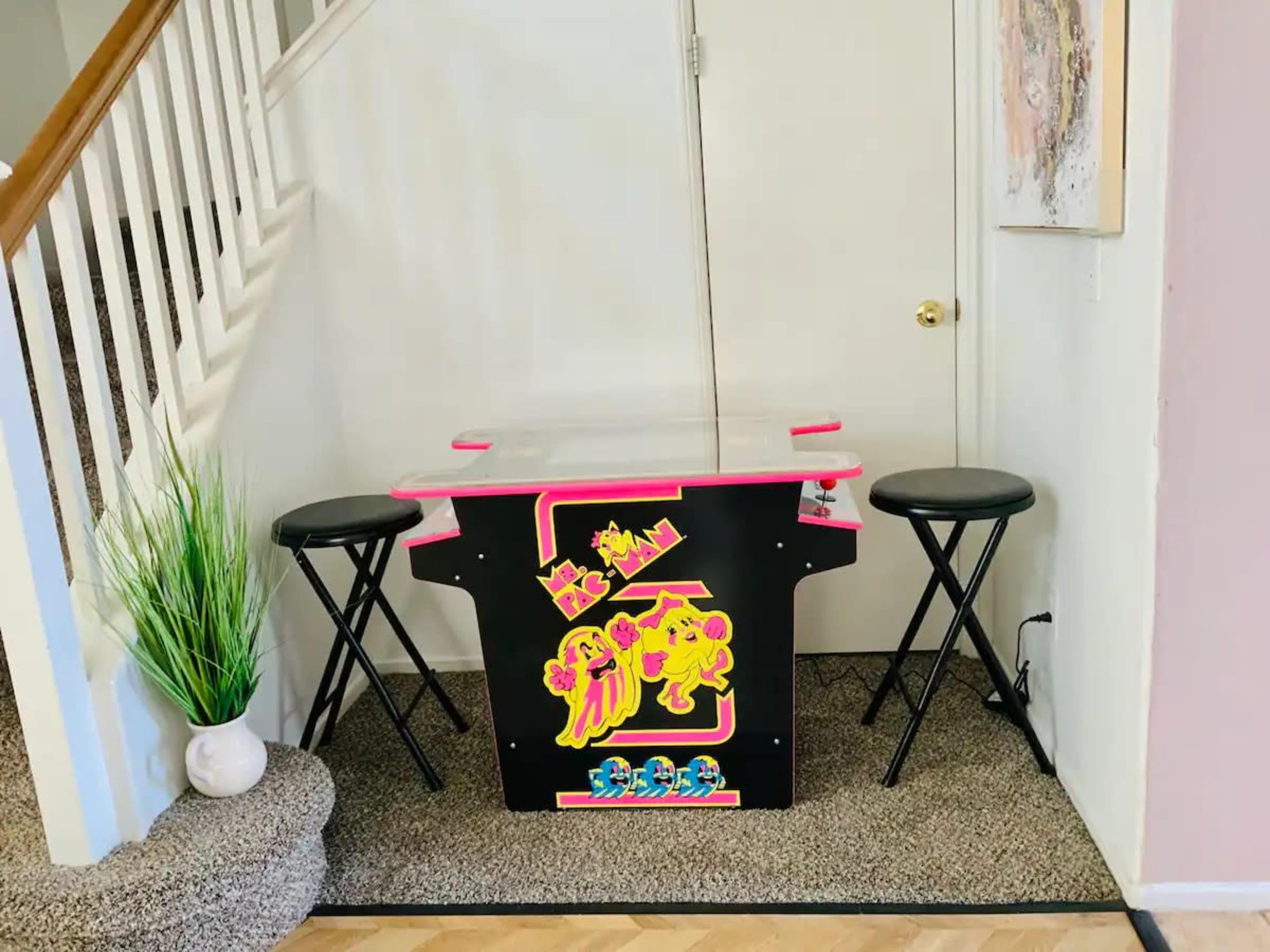A small arcade game table with stools is situated in a corner near a staircase, accompanied by a decorative plant.