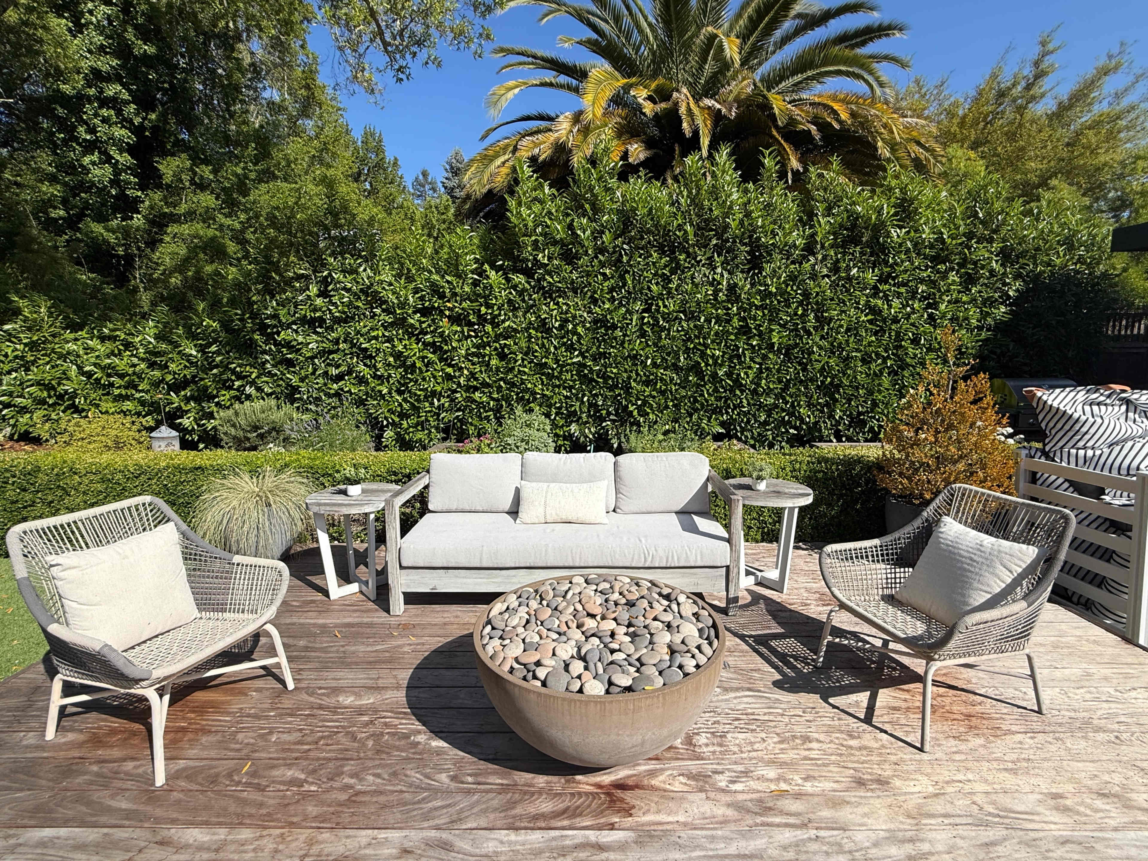 A seating area with a circular stone fire pit is surrounded by four chairs and a sofa on a wooden deck, with lush greenery and palm trees in the background.