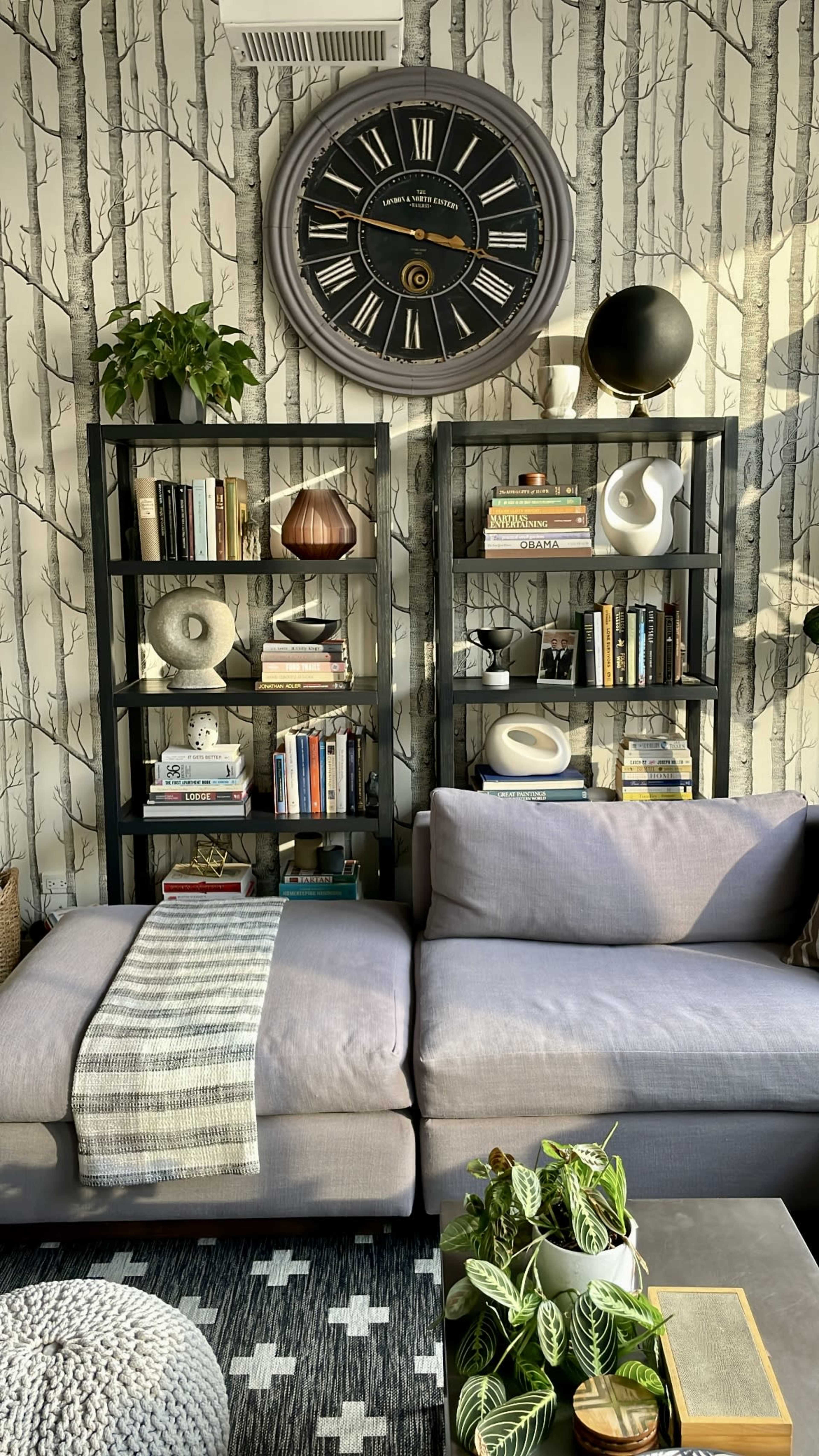 A modern living room features a gray sectional sofa, black bookshelves lined with books and decor, a round wall clock, and a coffee table with a plant, all set against a patterned wallpaper of trees.